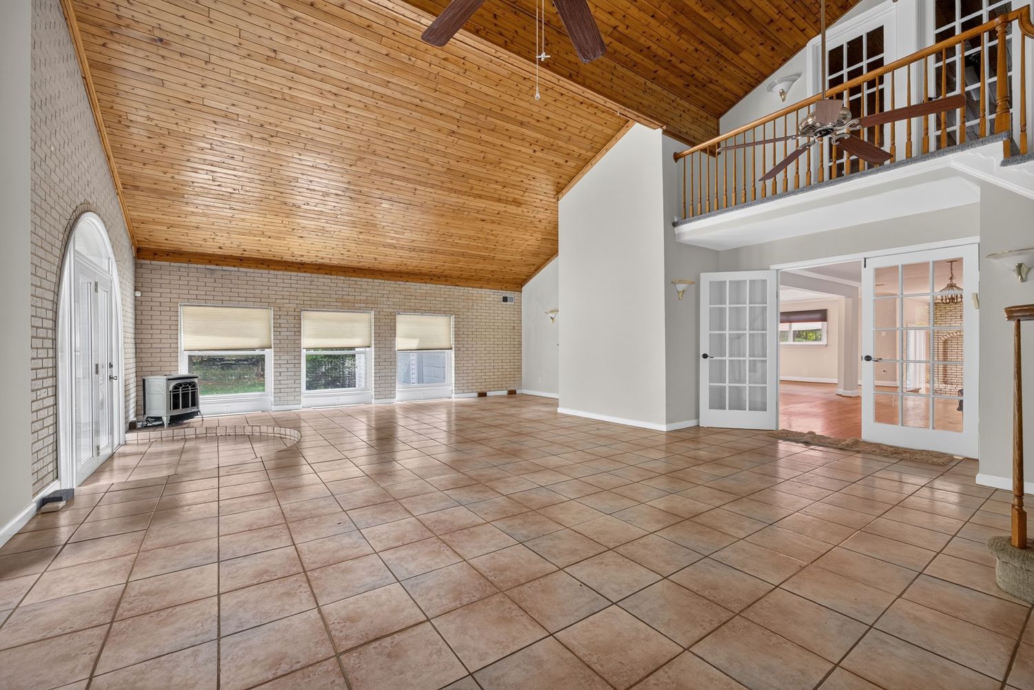 Spacious living room with high wood-paneled vaulted ceiling, tile floors, brick wall, ceiling fan, and open second-floor balcony.