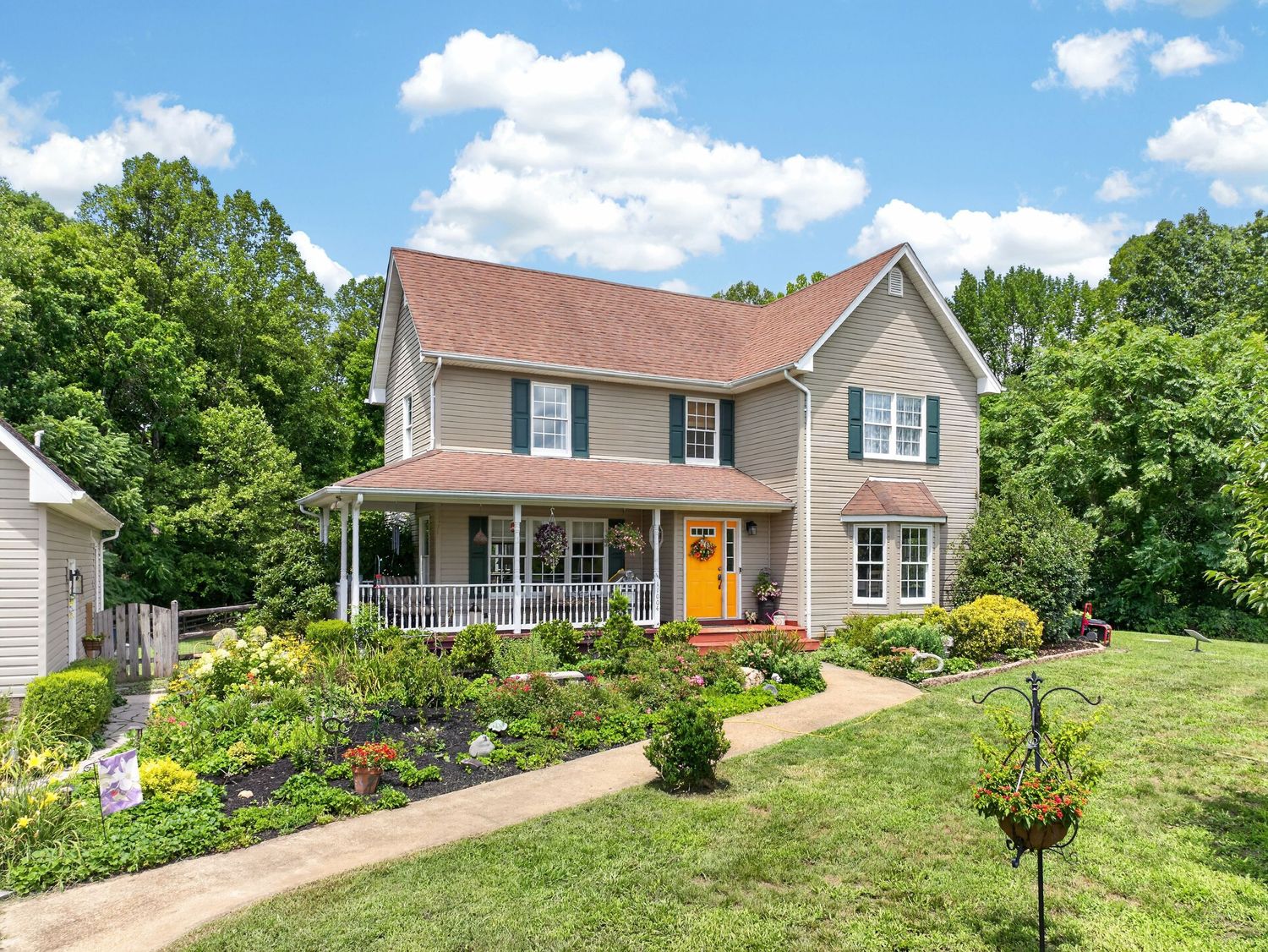 Two-story colonial home with covered porch, yellow front door, manicured gardens, and lush green trees in background.