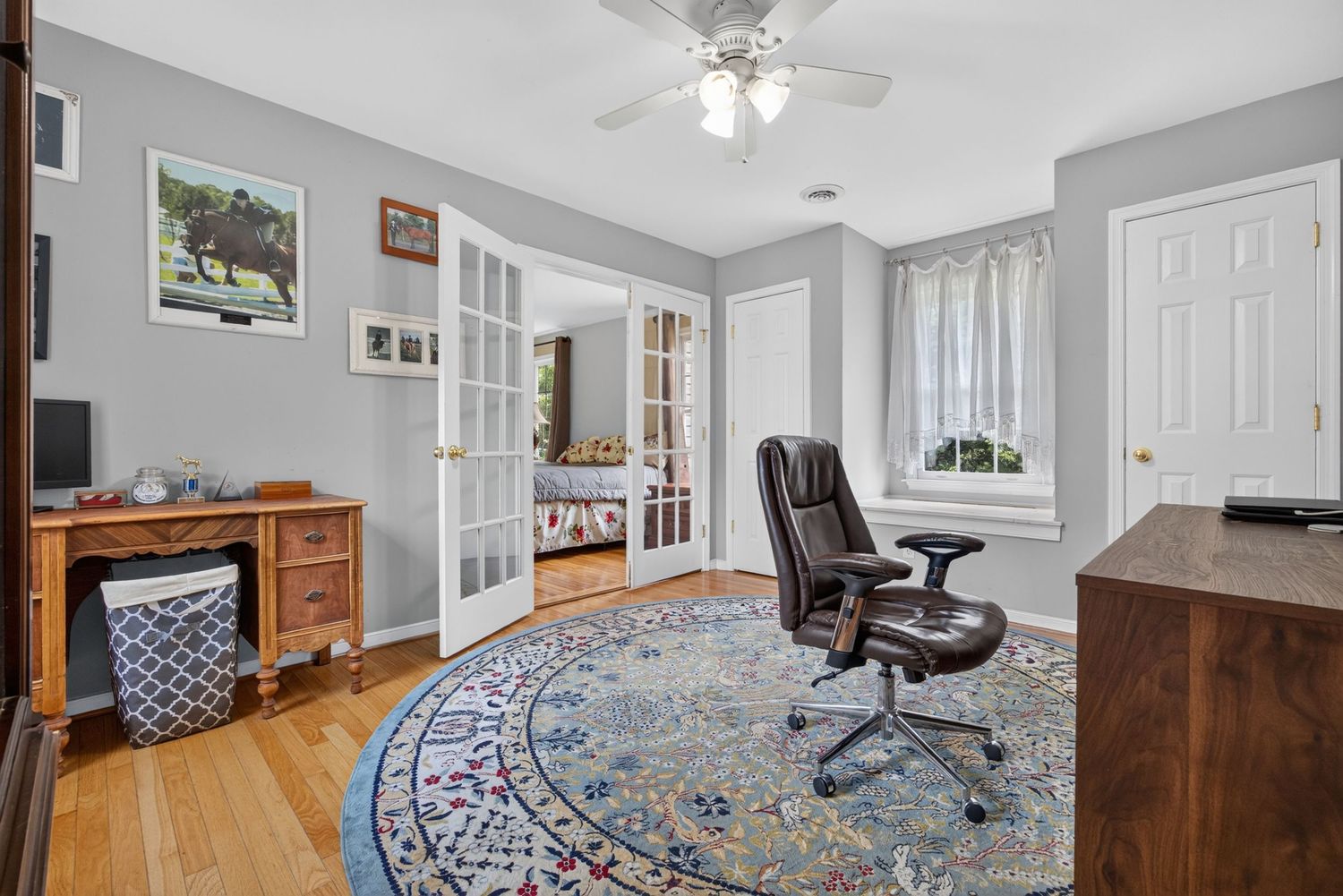 Home office with hardwood floors, blue floral rug, leather chair, French doors, ceiling fan, and natural light from window.