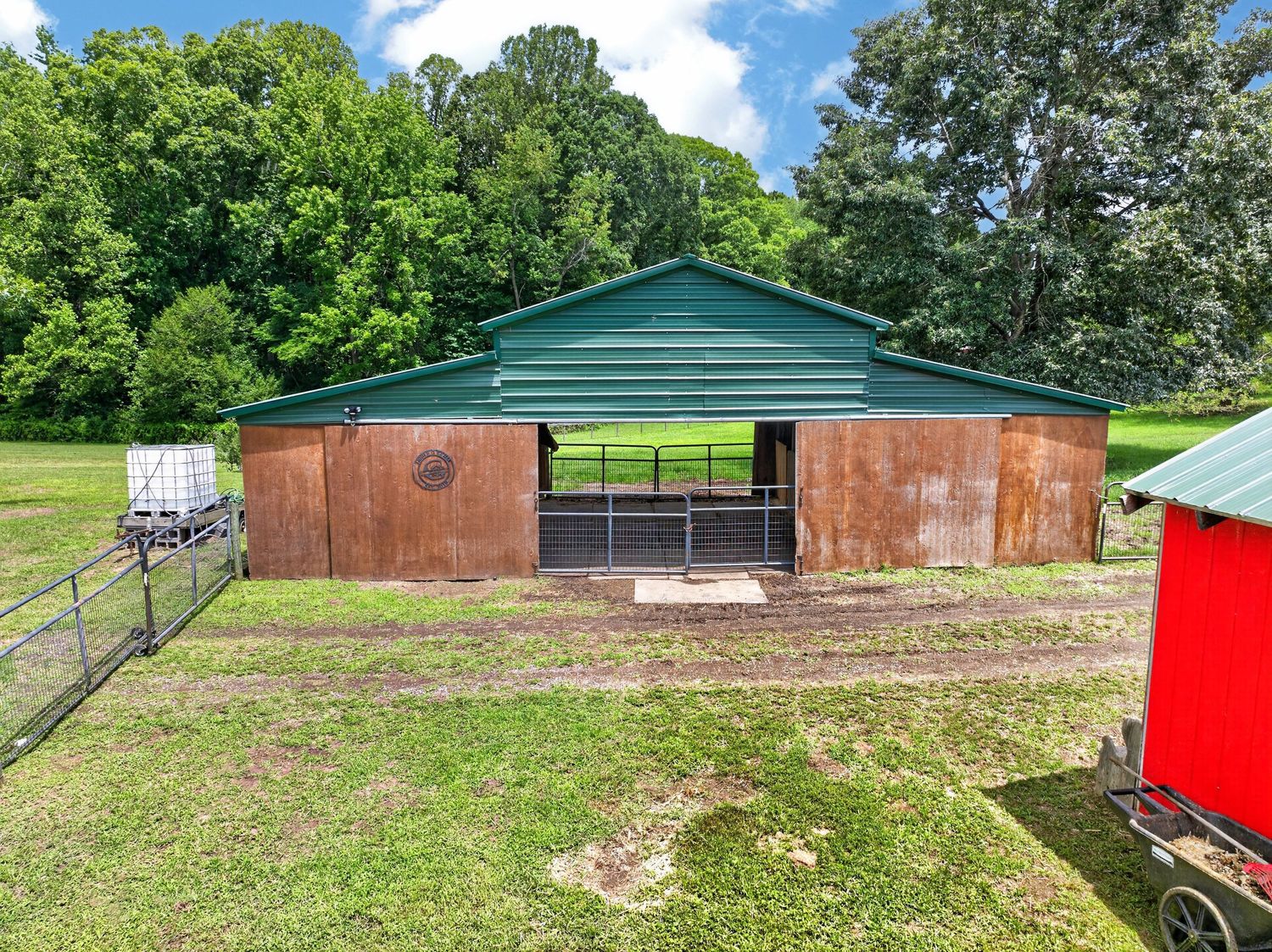 Green metal barn with wooden panels and open front on a rural property surrounded by lush trees and green grass.