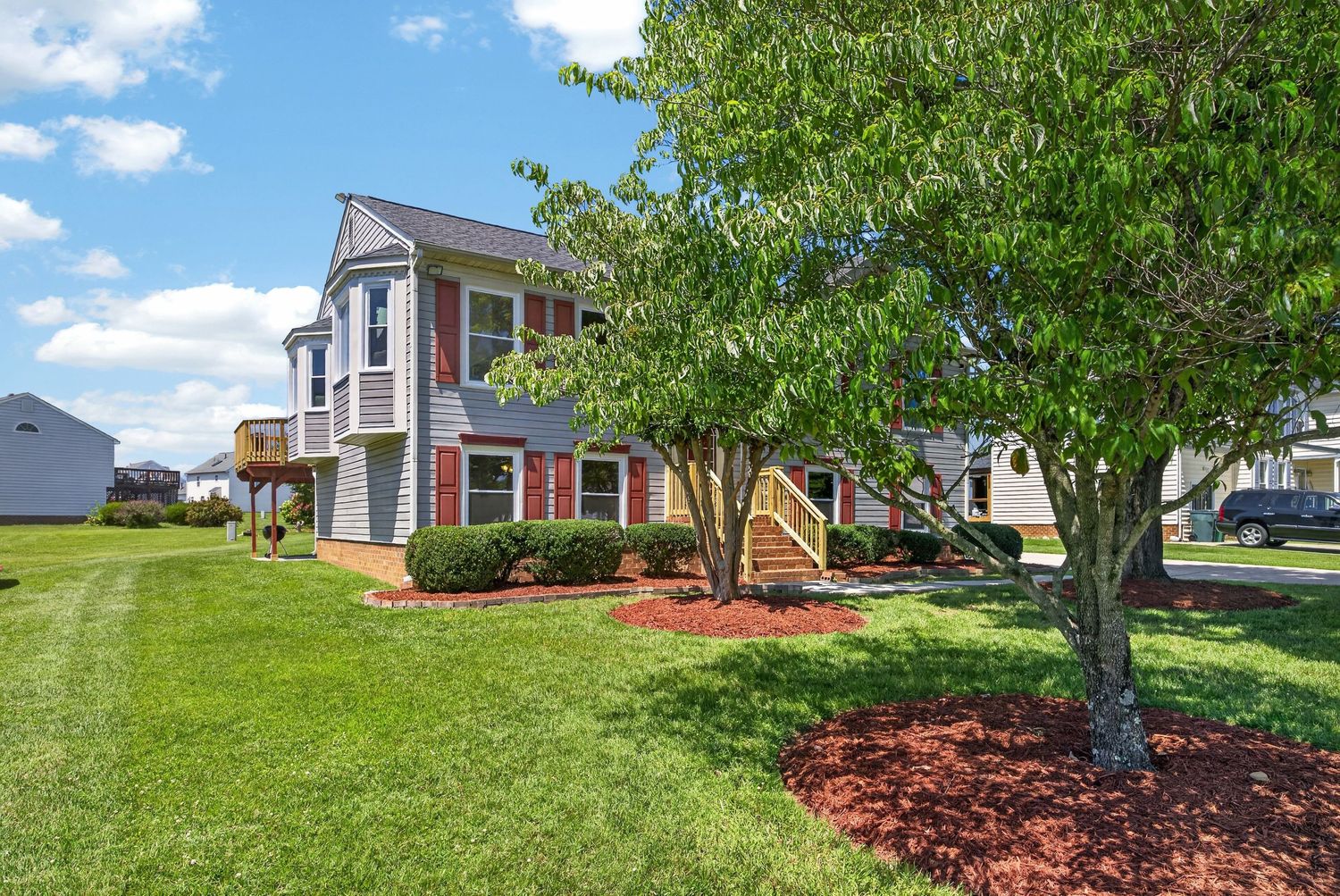Two-story residential home with gray siding, red brick accents, lush green lawn, and mature shade trees under a blue sky.