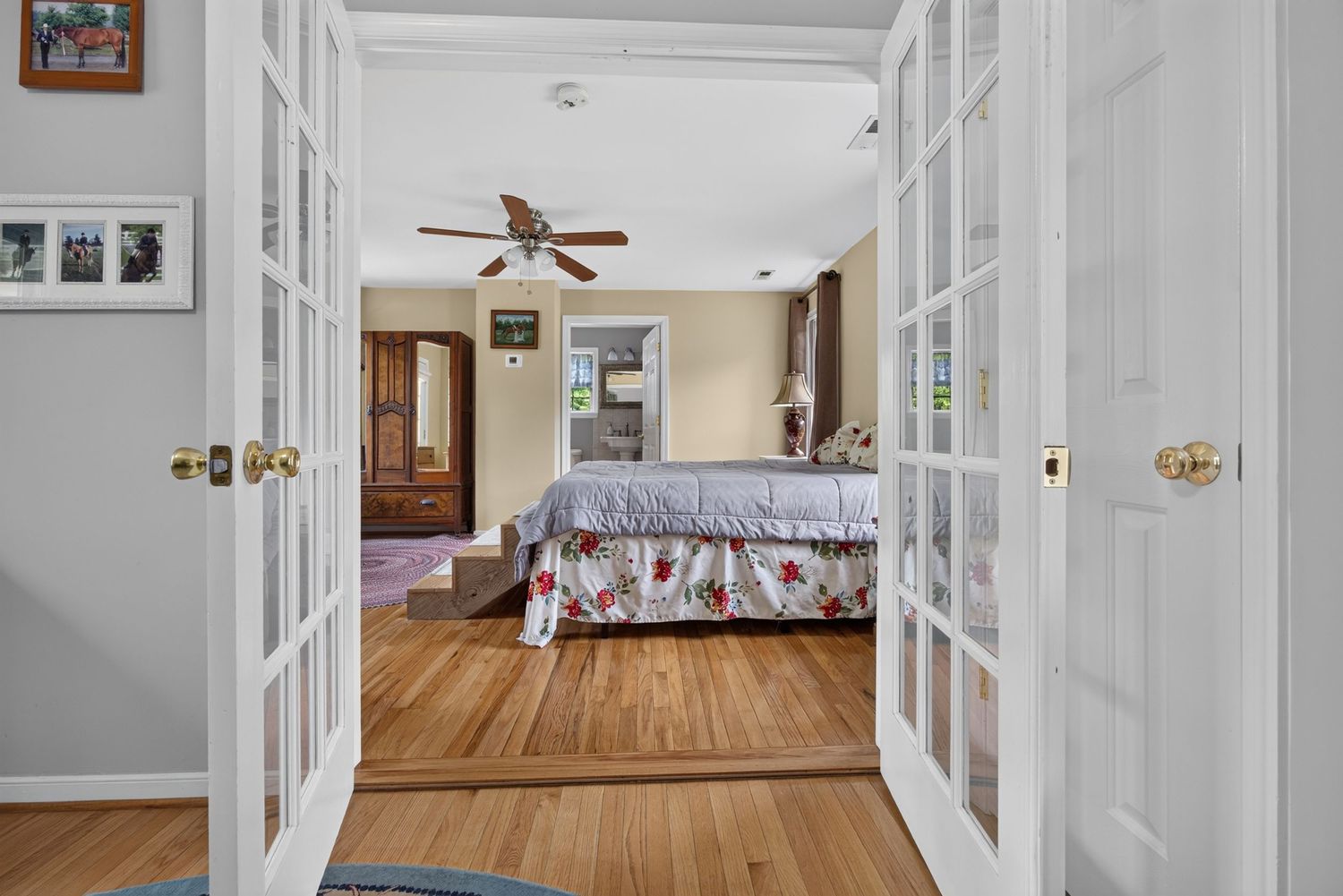 Elegant bedroom viewed through French doors featuring hardwood floors, ceiling fan, and natural light from multiple windows.