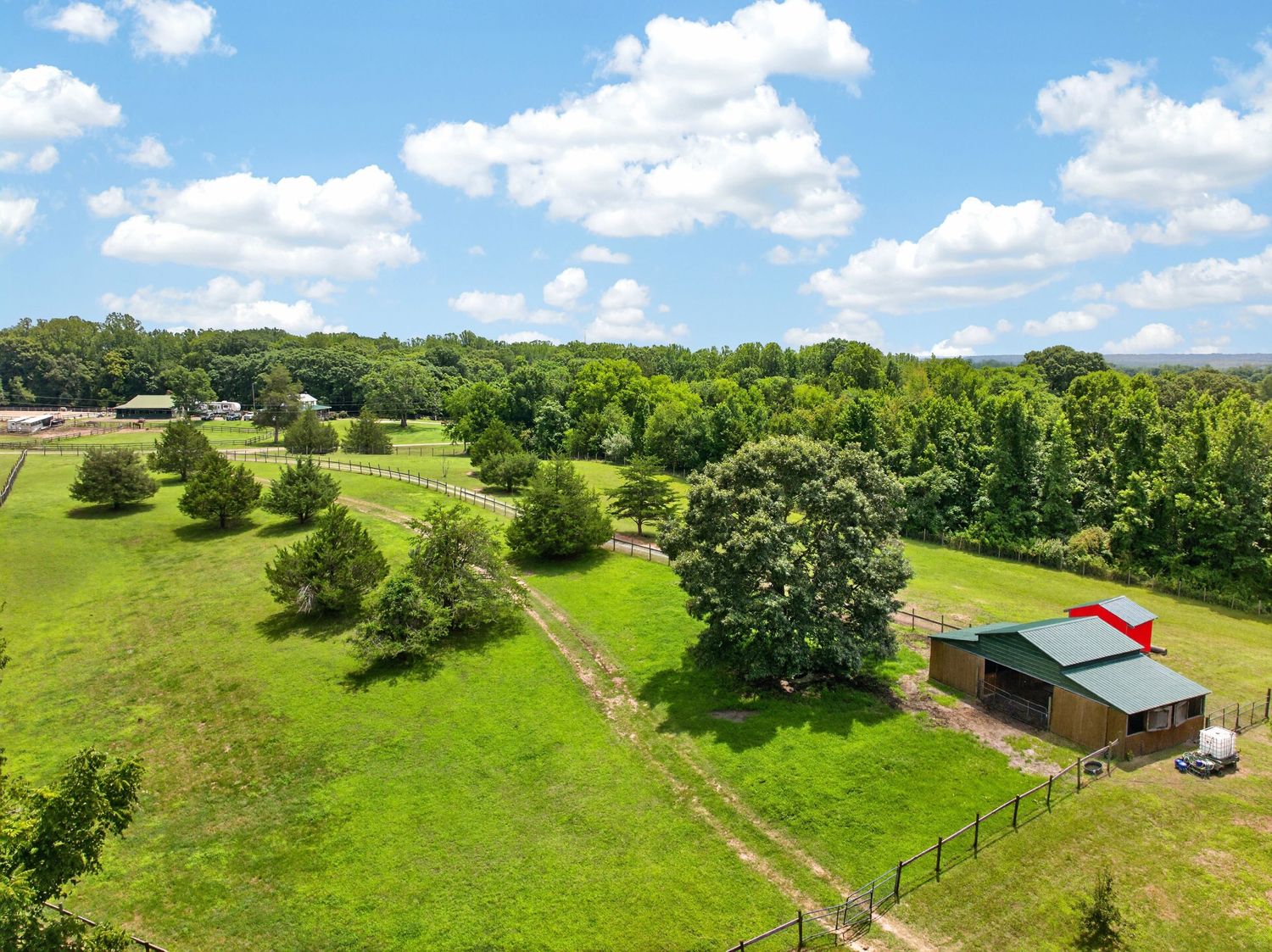 Aerial view of a lush green rural property featuring a red-roofed barn, open pastures, fenced areas, and mature trees.