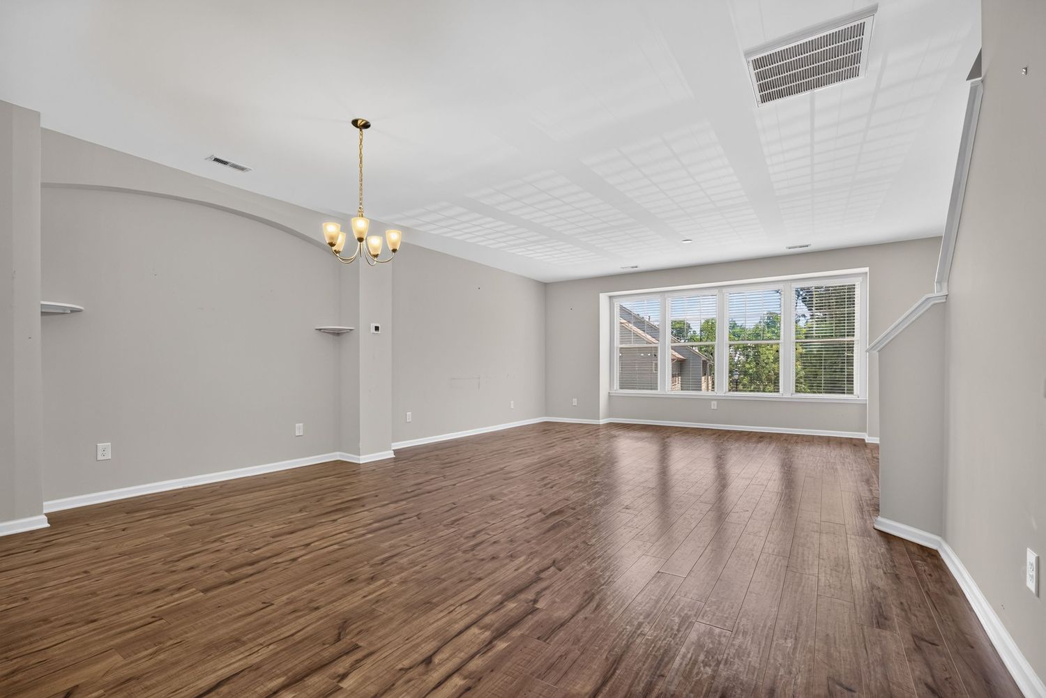 Spacious living room with dark hardwood floors, gray walls, chandelier, and bright window with white trim.