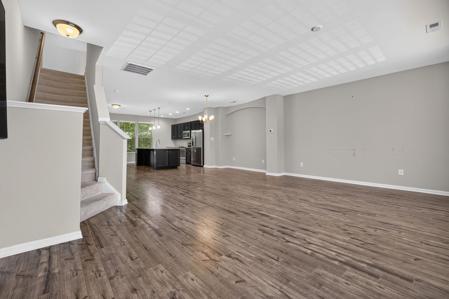Spacious open-concept living room with dark hardwood floors, neutral gray walls, staircase, and view into modern kitchen.