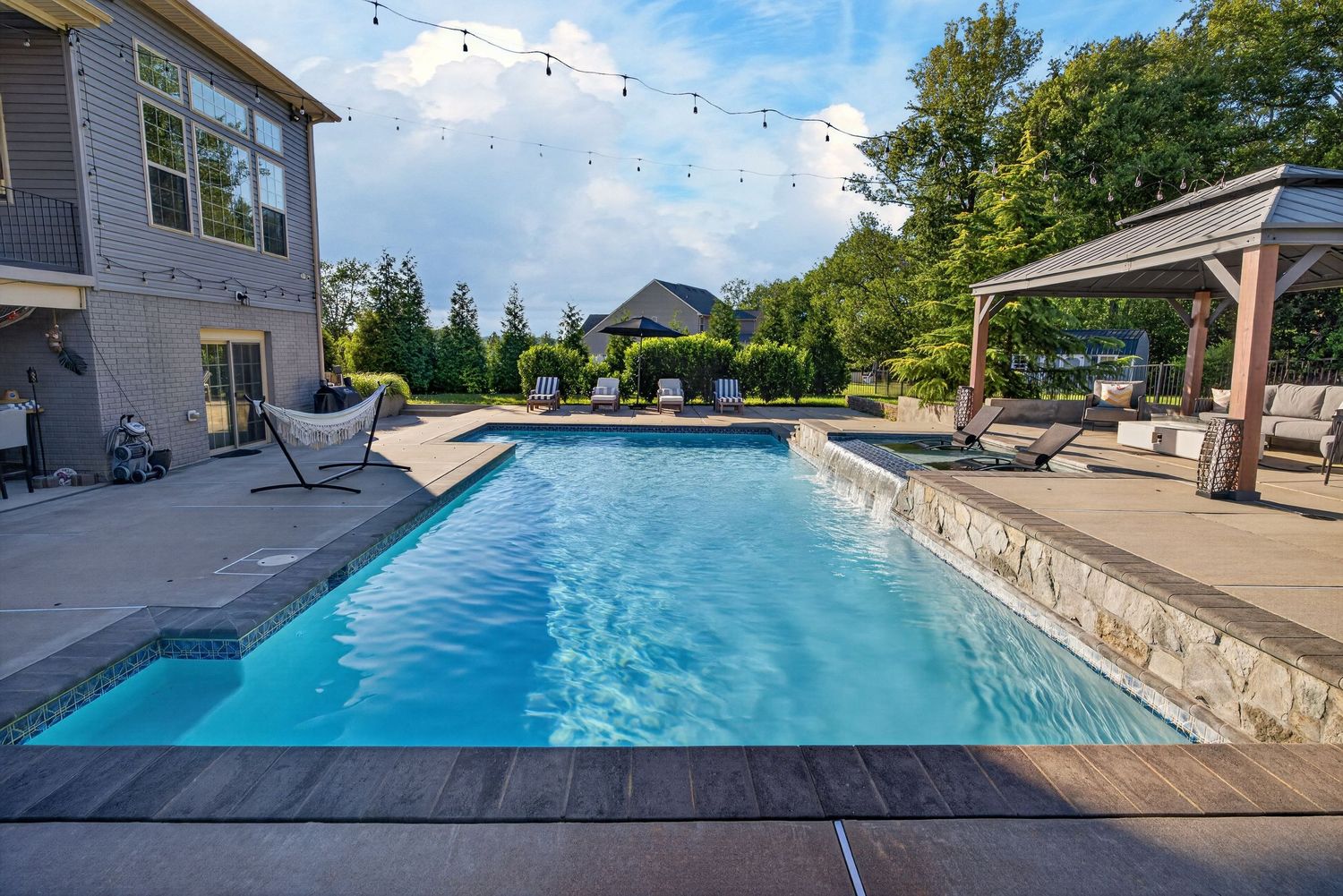 Luxurious backyard pool with stone patio, pergola, lounge chairs, and string lights surrounded by lush trees.