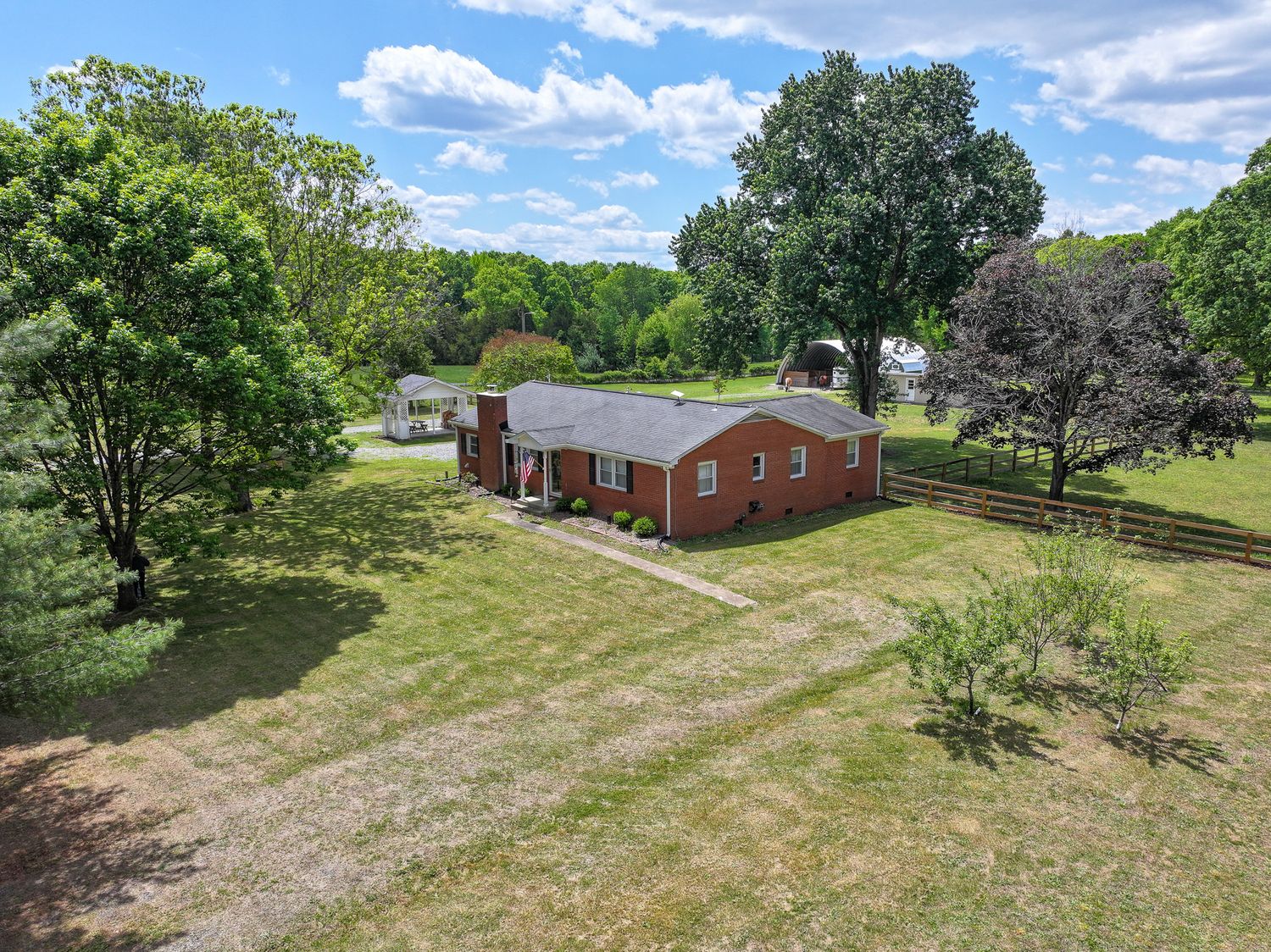Aerial view of a red brick ranch home surrounded by lush green trees and a spacious yard with a wooden fence.
