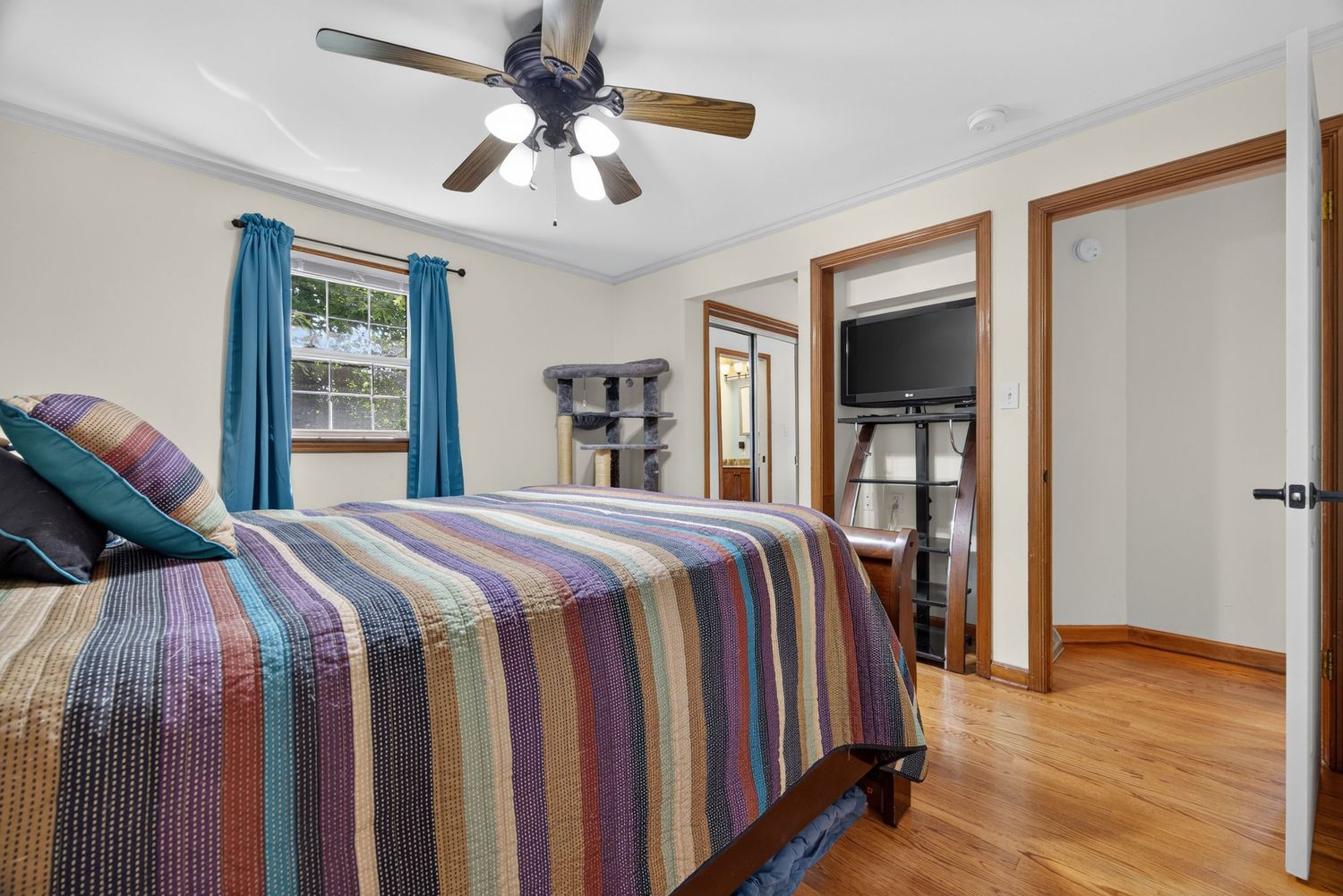 Bright bedroom with striped quilt, ceiling fan, hardwood floors, and open doors leading to a sunlit sitting area with TV.