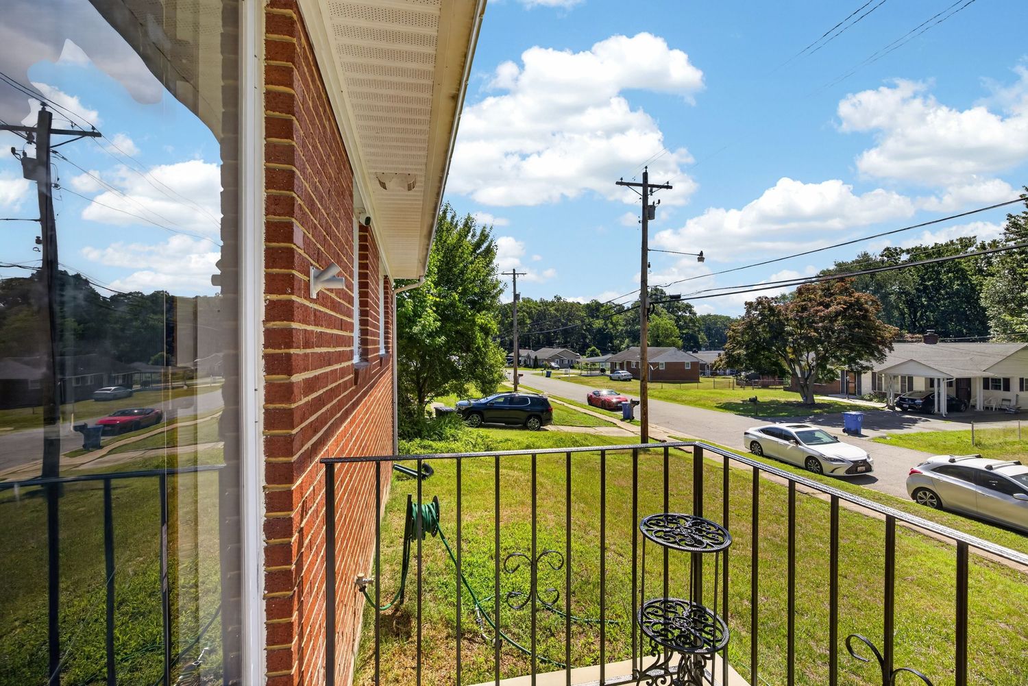 Brick home balcony overlooking a green lawn and quiet suburban street on a sunny day with blue skies.