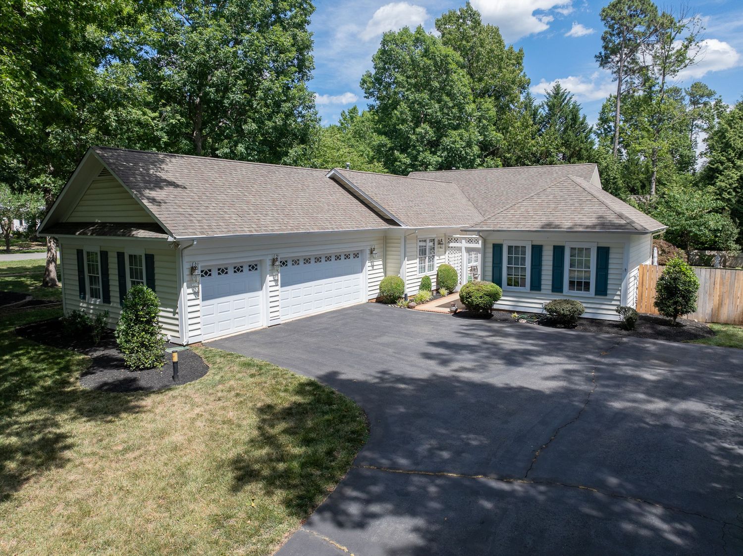 Aerial view of a spacious ranch-style home with two-car garage, blue shutters, and lush green trees in a suburban setting.