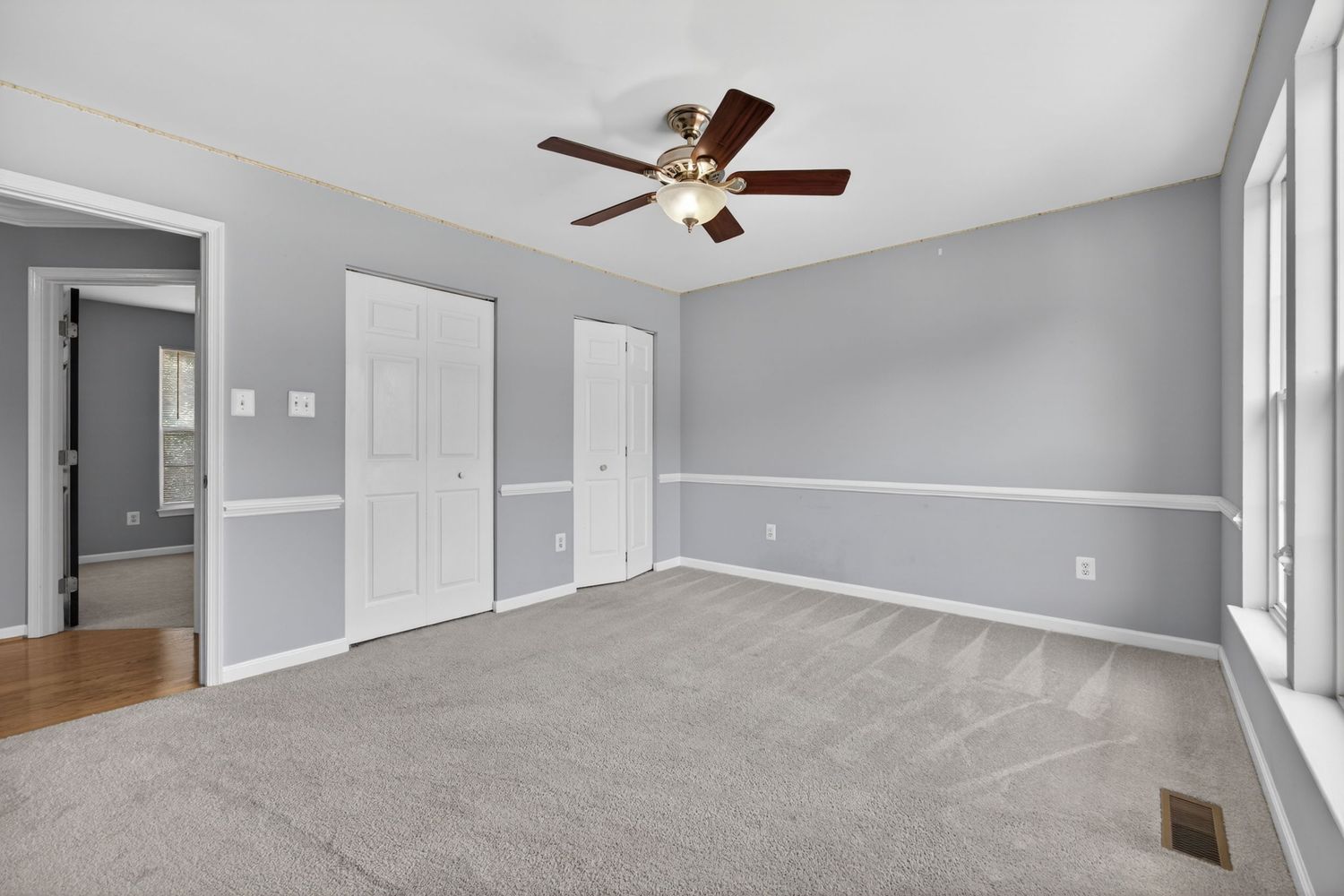 Spacious bedroom with gray carpet, white wainscoting, ceiling fan, two closets, and neutral gray walls in a modern home.