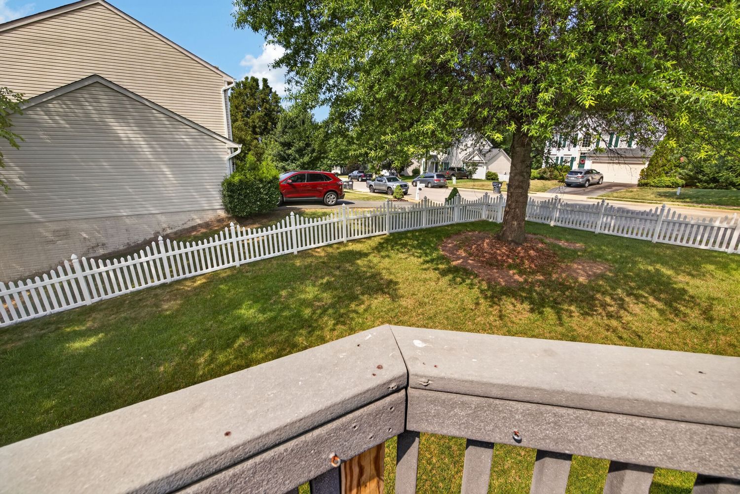 Wooden deck overlooking a lush green yard with a white picket fence, large trees, and a red car parked nearby.