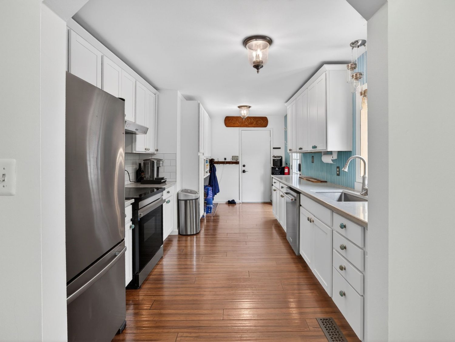 Modern galley kitchen with white cabinets, stainless steel appliances, hardwood floors, and blue tile backsplash.