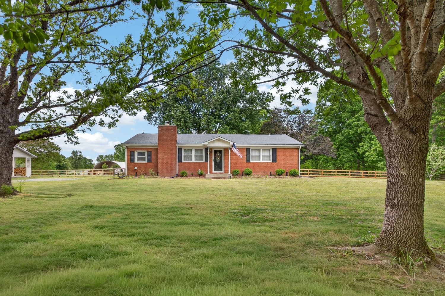 Red brick ranch home with chimney sits on lush green lawn surrounded by mature trees on a sunny day.