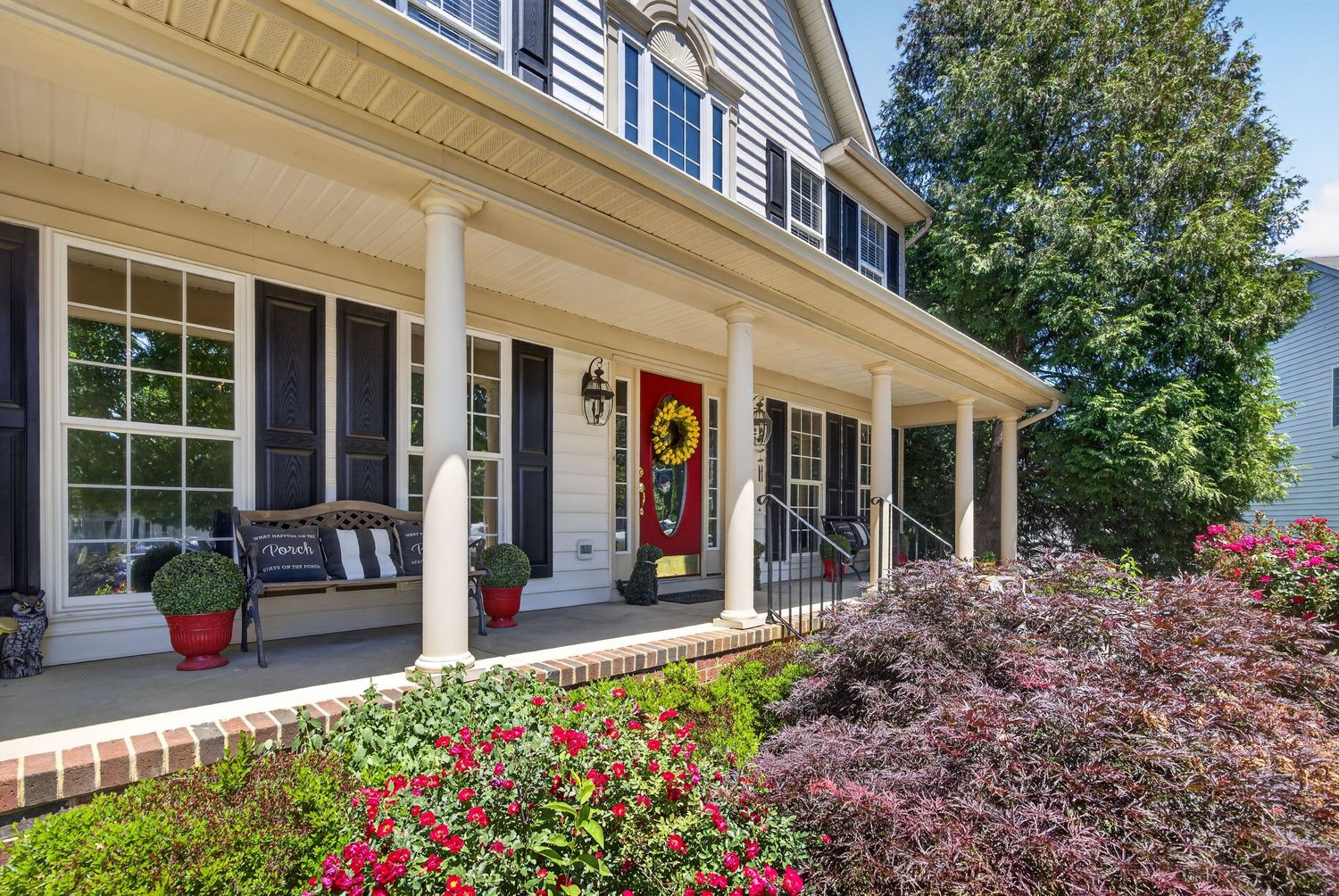 Charming colonial home with white columns, wraparound porch, red front door, and vibrant flower gardens in full bloom.