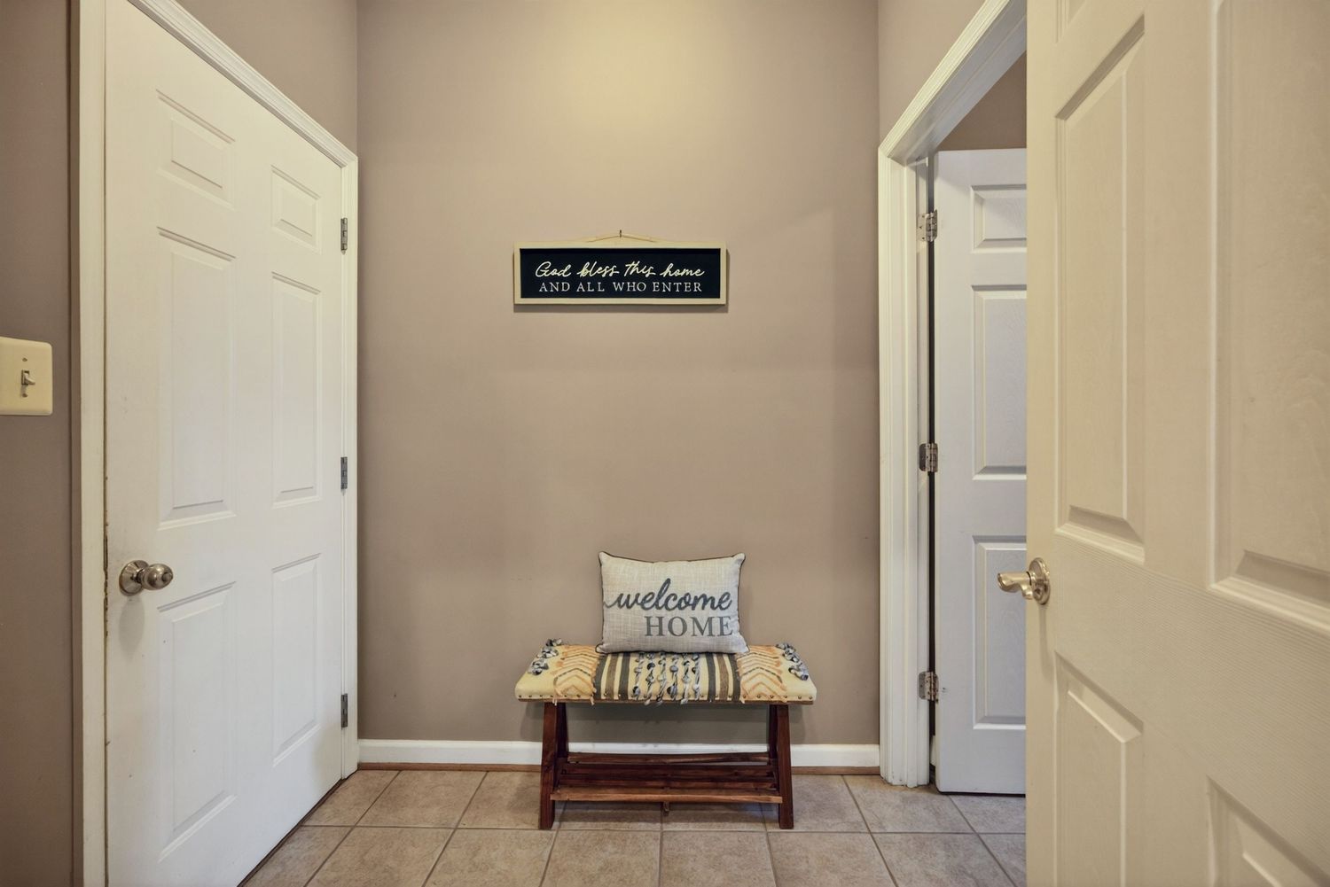 Cozy home entryway with welcome bench, decorative sign, white doors, and neutral beige walls on tile flooring.