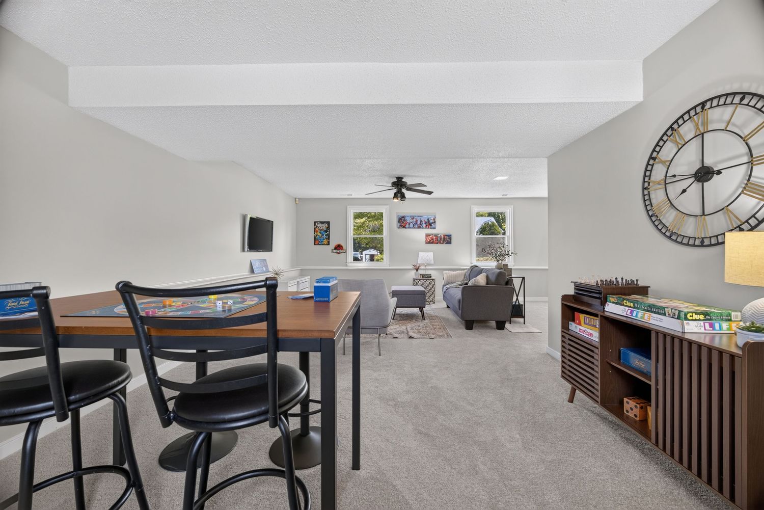 Spacious living area with carpet, bar-height table, ceiling fan, large clock, and bookshelf filled with board games.