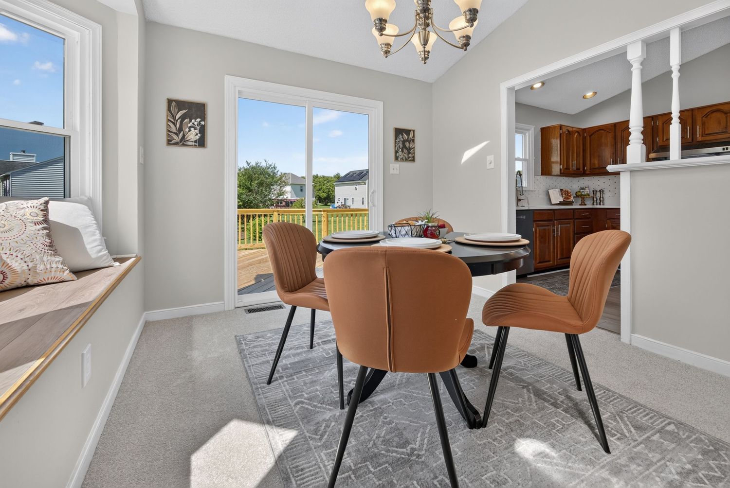 Bright dining room with tan chairs, round table, chandelier, patterned rug, and sliding glass door to deck.