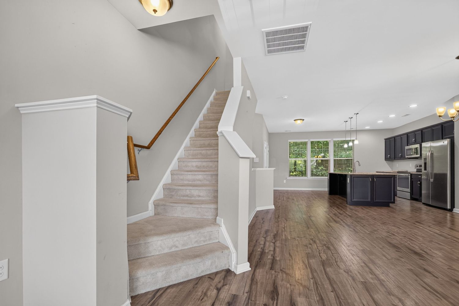 Open-concept home interior featuring hardwood floors, white staircase with wood railing, and modern kitchen with dark cabinets.