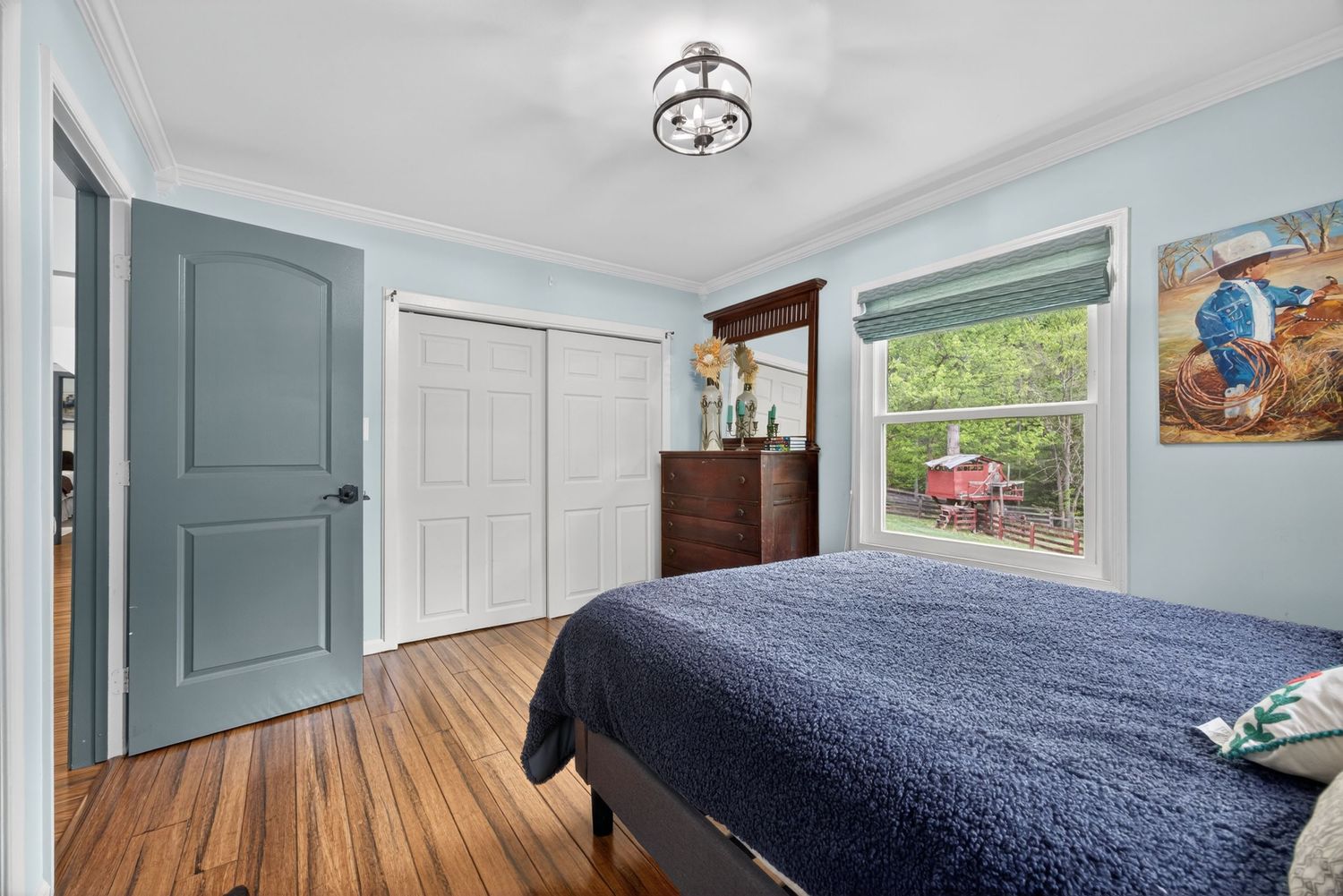 Cozy bedroom with hardwood floors, crown molding, closet, dresser, and a window with natural light and outdoor views.
