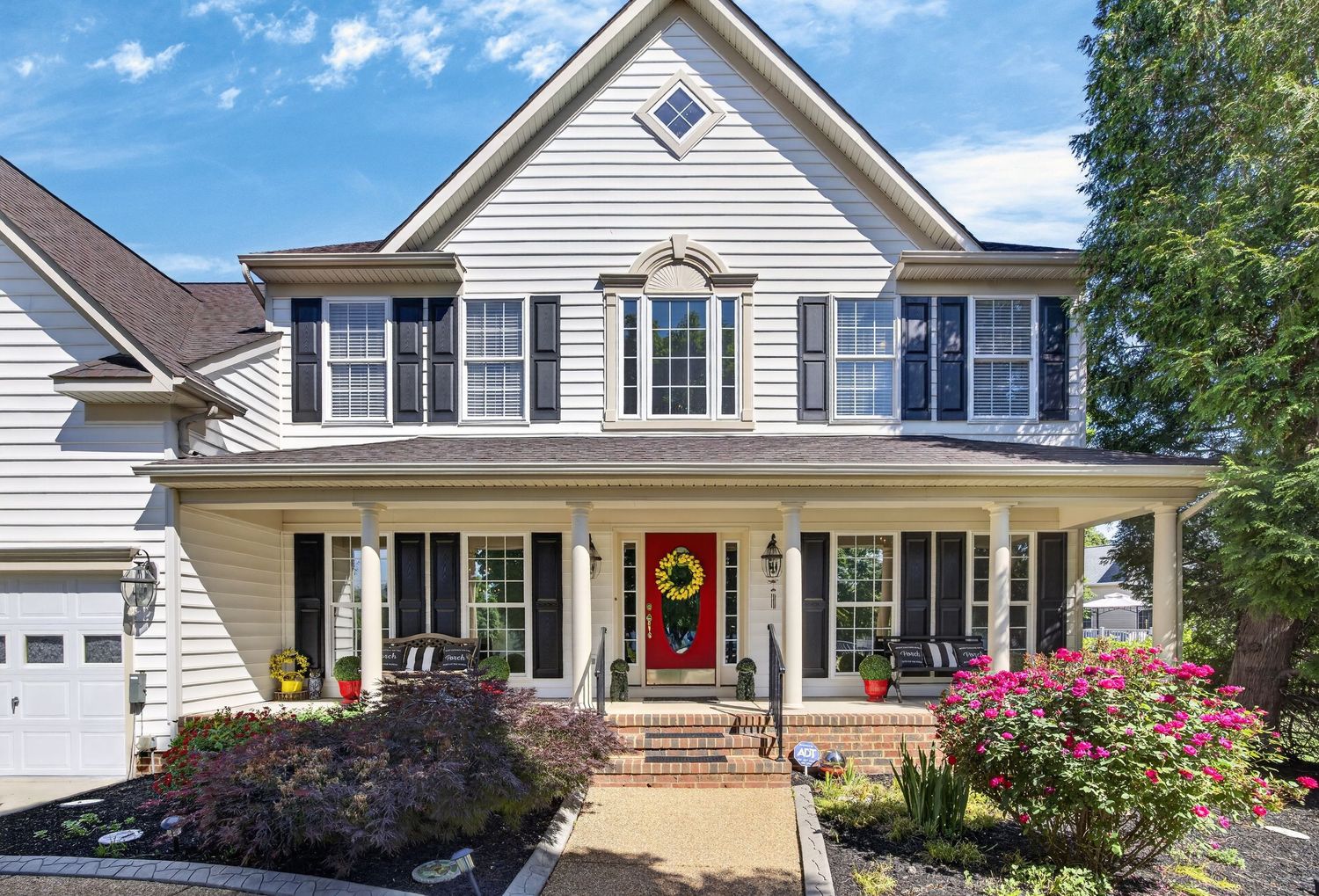White two-story colonial home with black shutters, red front door, covered porch, and blooming flowers in a manicured landscape.