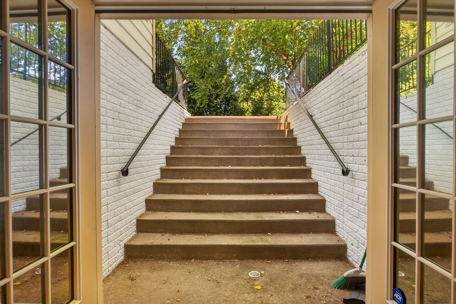 Outdoor concrete staircase with white brick walls, metal handrails, and lush green trees visible at the top.