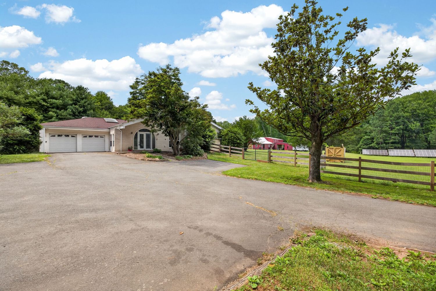A spacious paved driveway leads to a ranch-style home with a garage, wooden fence, and lush green trees under a blue sky.