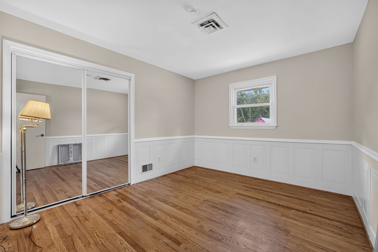 Bright empty bedroom with hardwood floors, white wainscoting, mirrored closet doors, and a window with natural light.