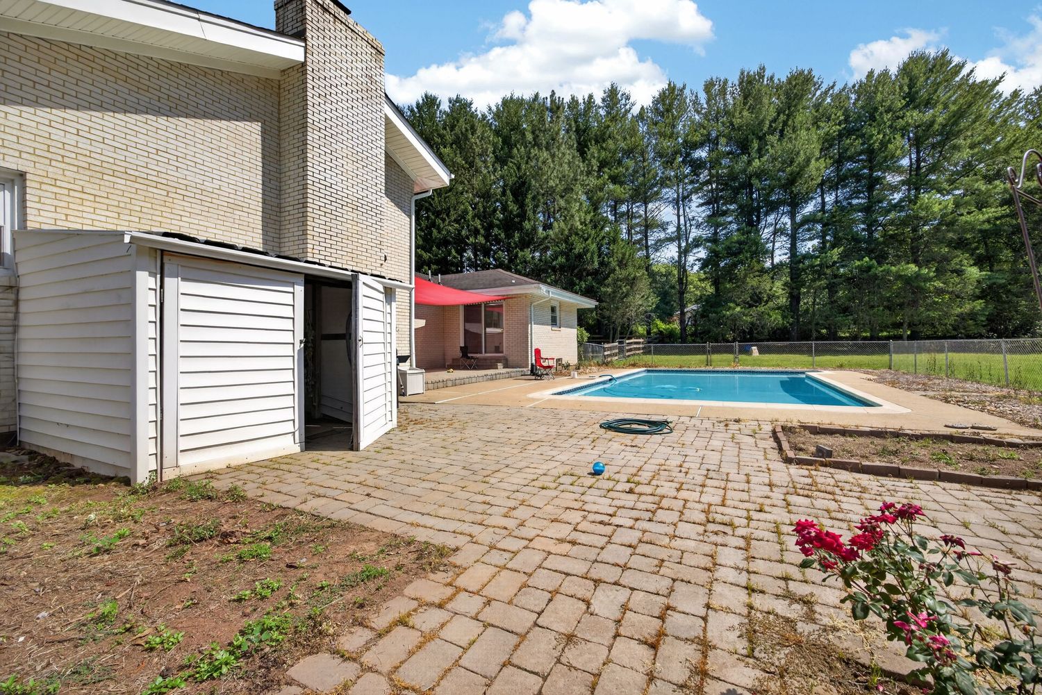 Brick patio with pool, shed, and lush green trees in a spacious backyard on a sunny day.
