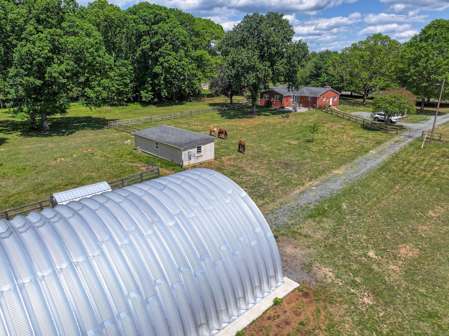 Aerial view of a rural farm property with a metal Quonset hut, grazing horses, red barn, and lush green trees.