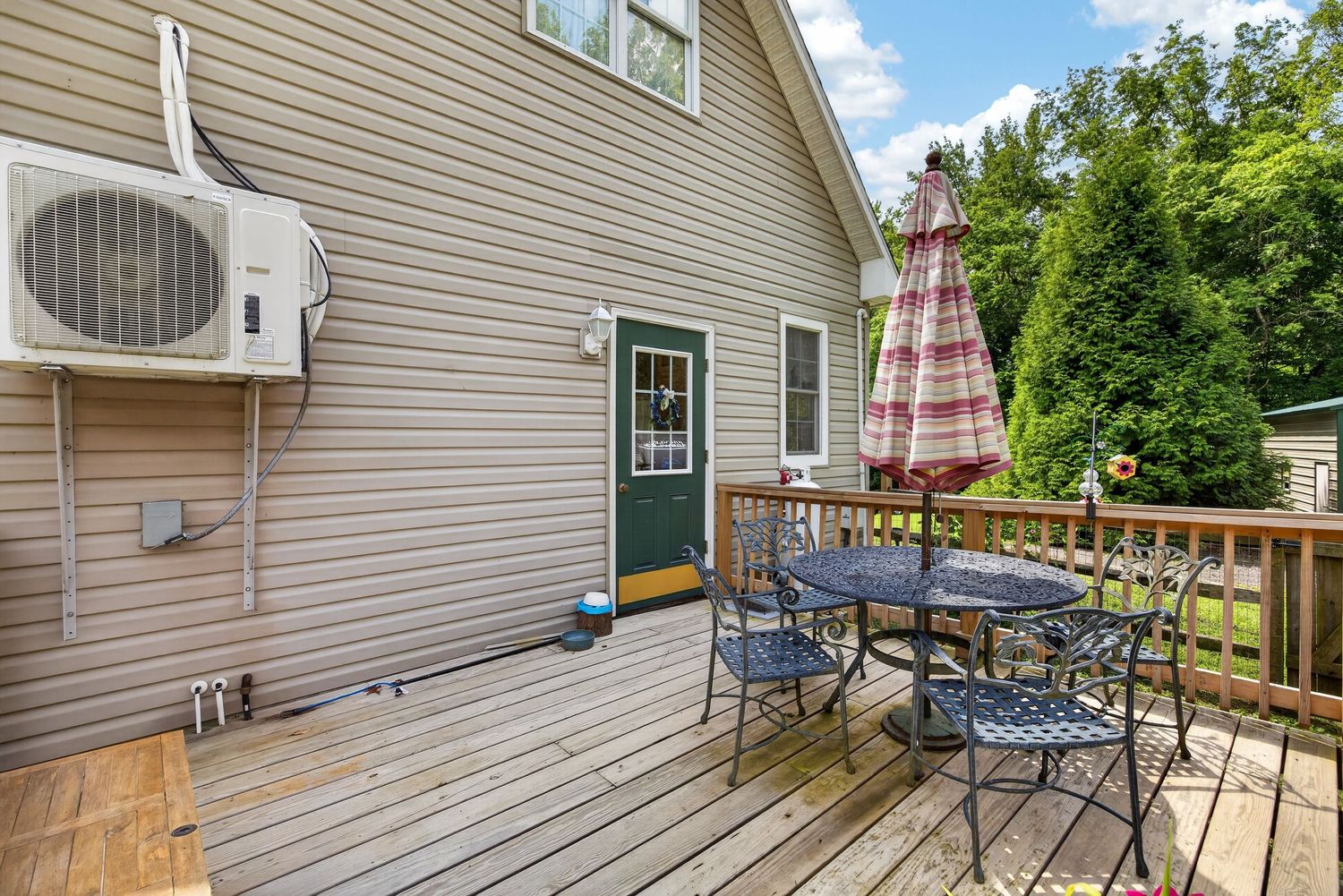Wooden deck with patio table, chairs, and umbrella beside a house with a mini-split AC unit and green entry door.