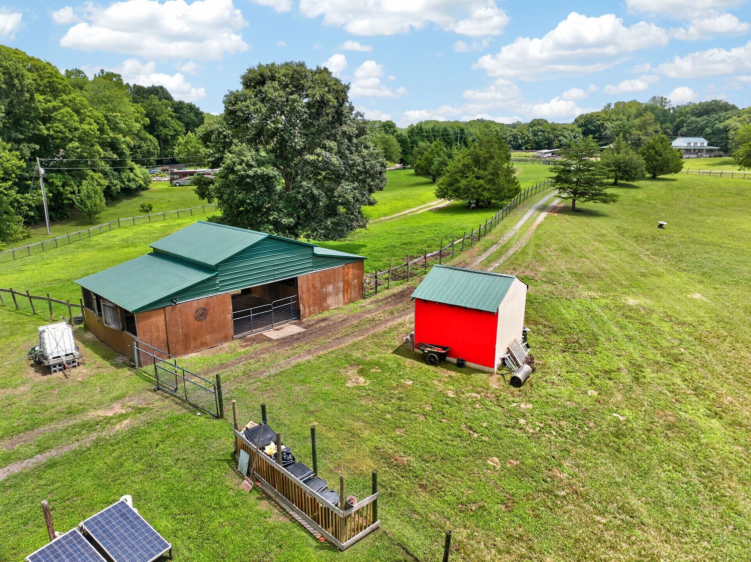 Aerial view of a rural farm property featuring a green-roofed barn, red shed, fenced pastures, and lush green trees.