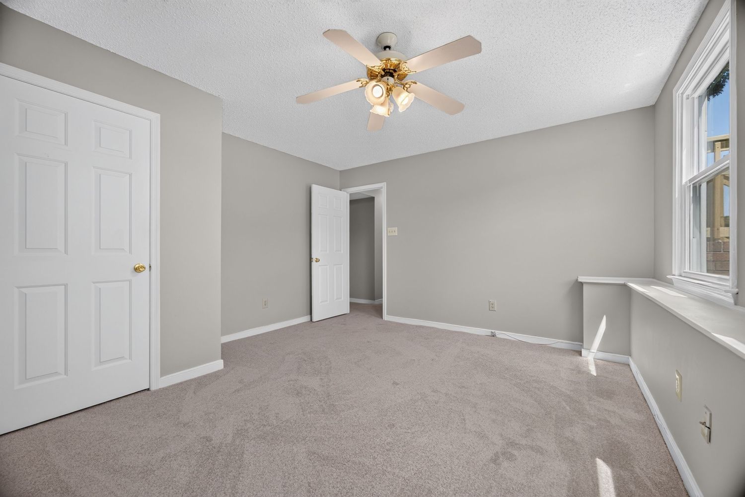 Spacious bedroom with gray carpet, ceiling fan, white doors, and natural light from a window.