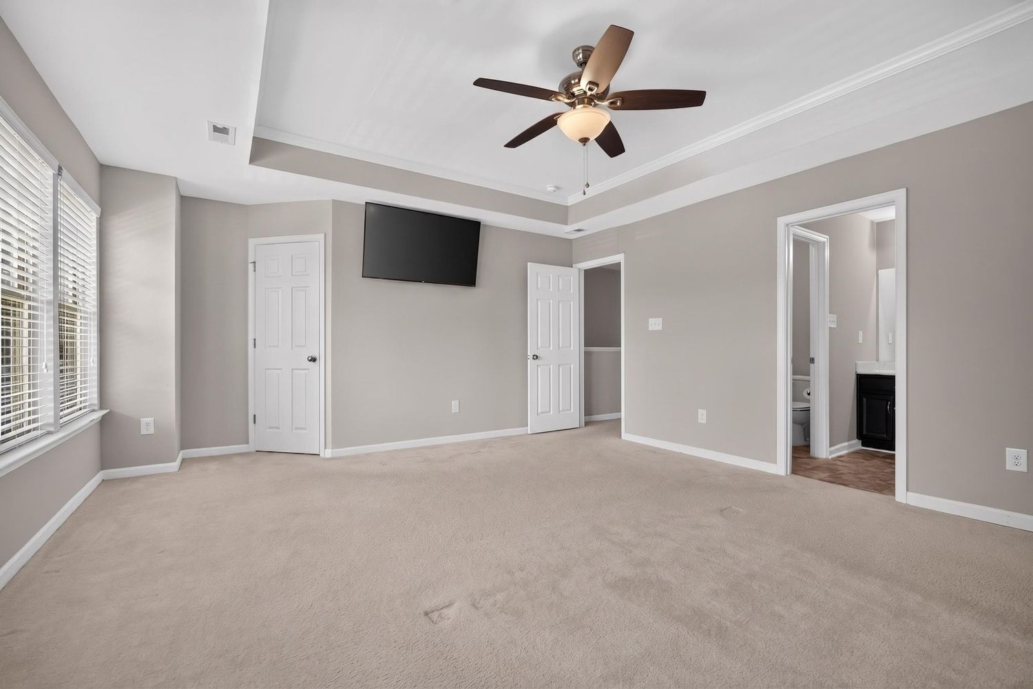 Spacious master bedroom with tray ceiling, ceiling fan, neutral carpet, wall-mounted TV, and doors leading to ensuite bath.
