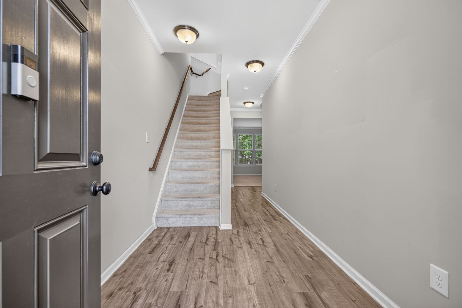 Bright entryway with light hardwood floors, white staircase, crown molding, and neutral walls leading to a window.
