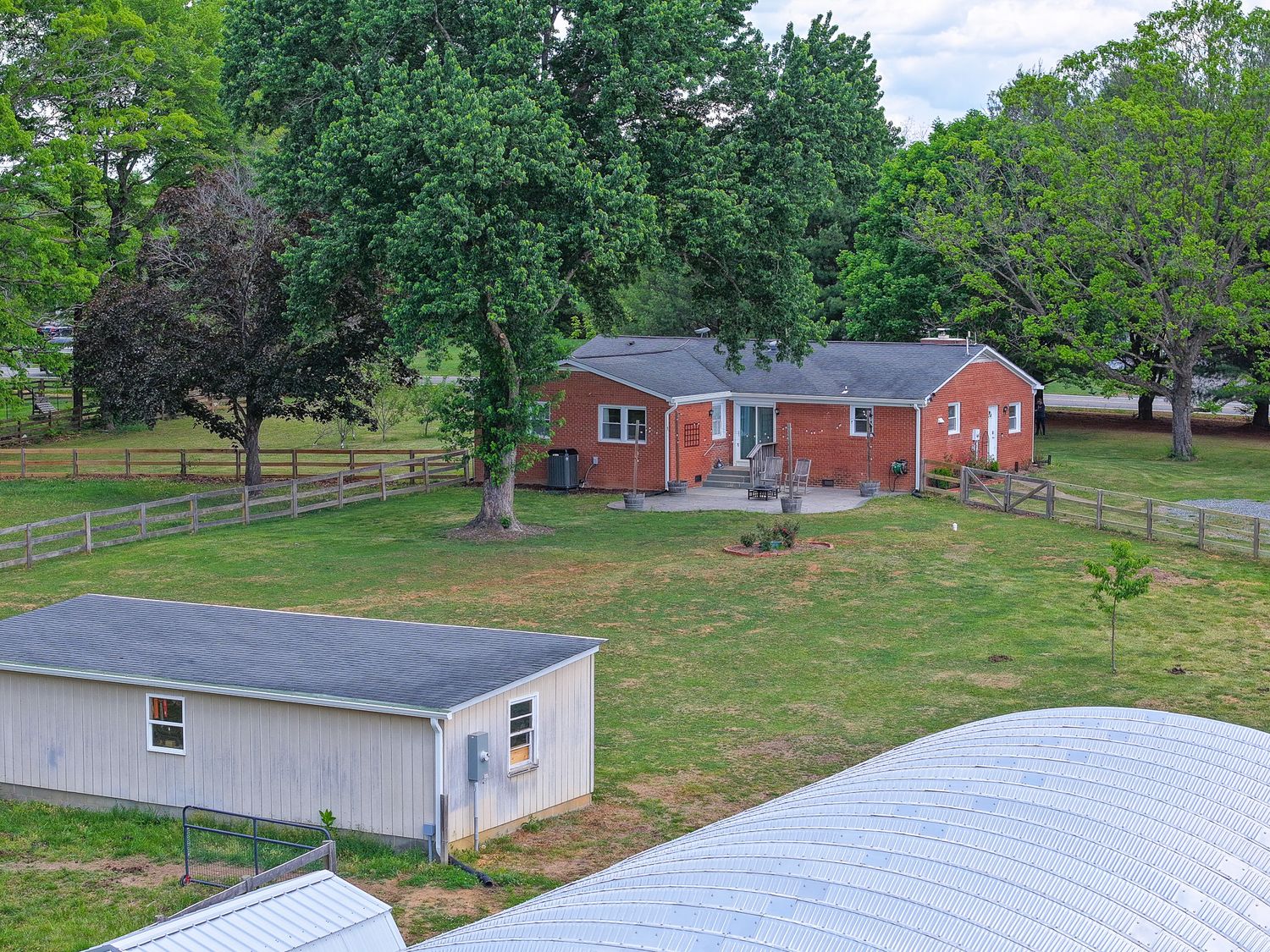 Aerial view of a brick ranch home with a gray roof, surrounded by lush green trees, fenced yard, and a small outbuilding.