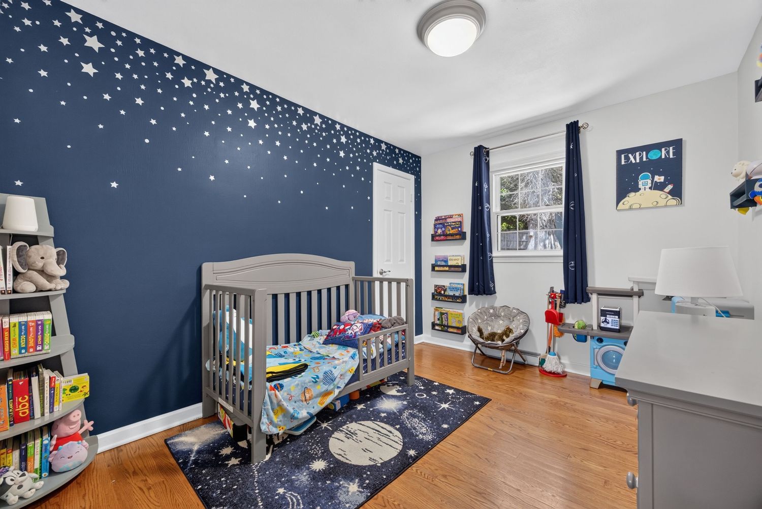 Space-themed nursery with navy star wall, gray crib, hardwood floors, and colorful galaxy rug in a bright, decorated room.