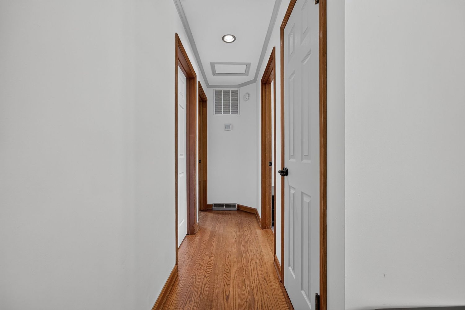 Narrow residential hallway with white walls, brown trim doors, hardwood floors, and recessed ceiling light.