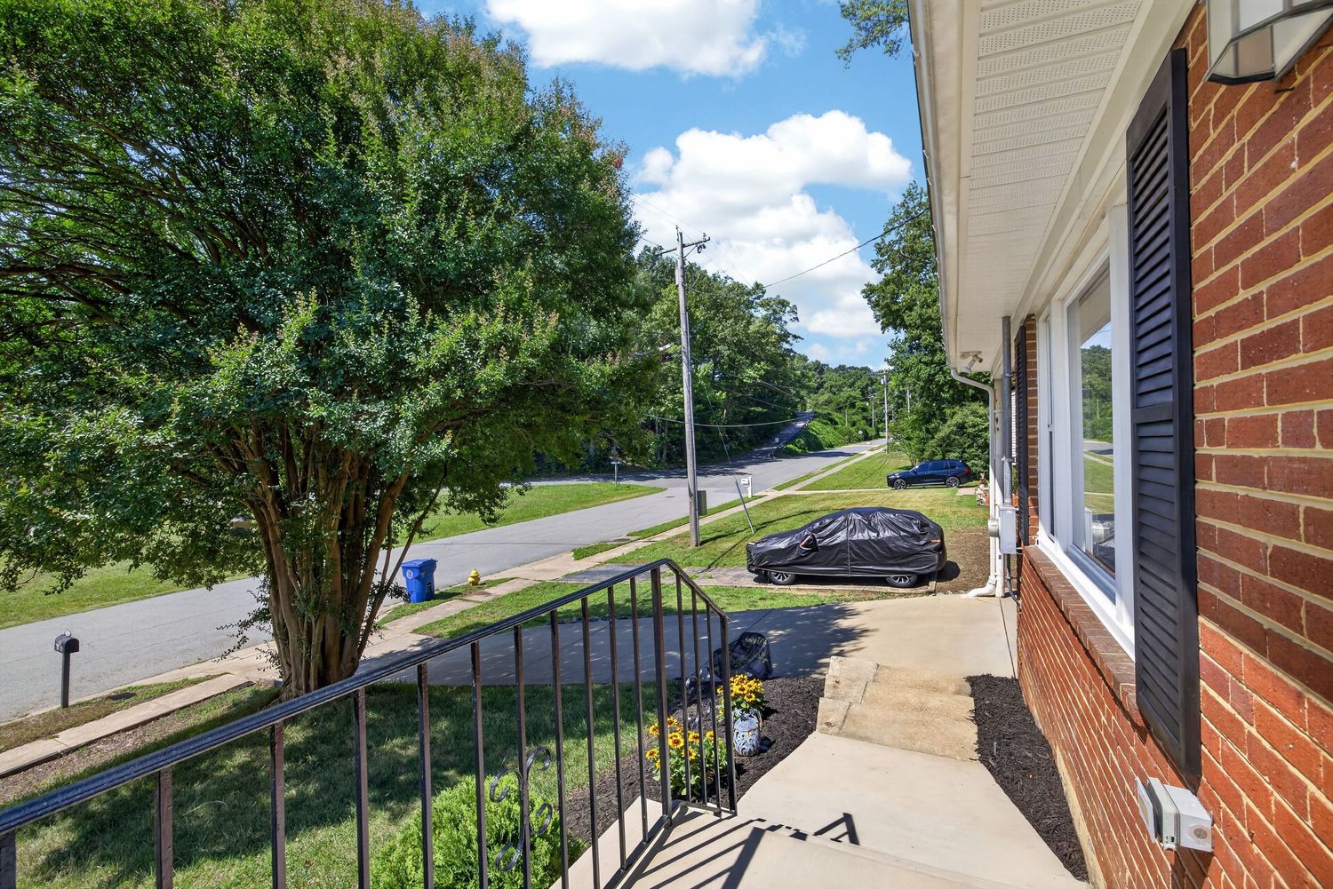 Brick home exterior featuring a front patio, black metal railing, mature trees, and a quiet suburban street on a sunny day.