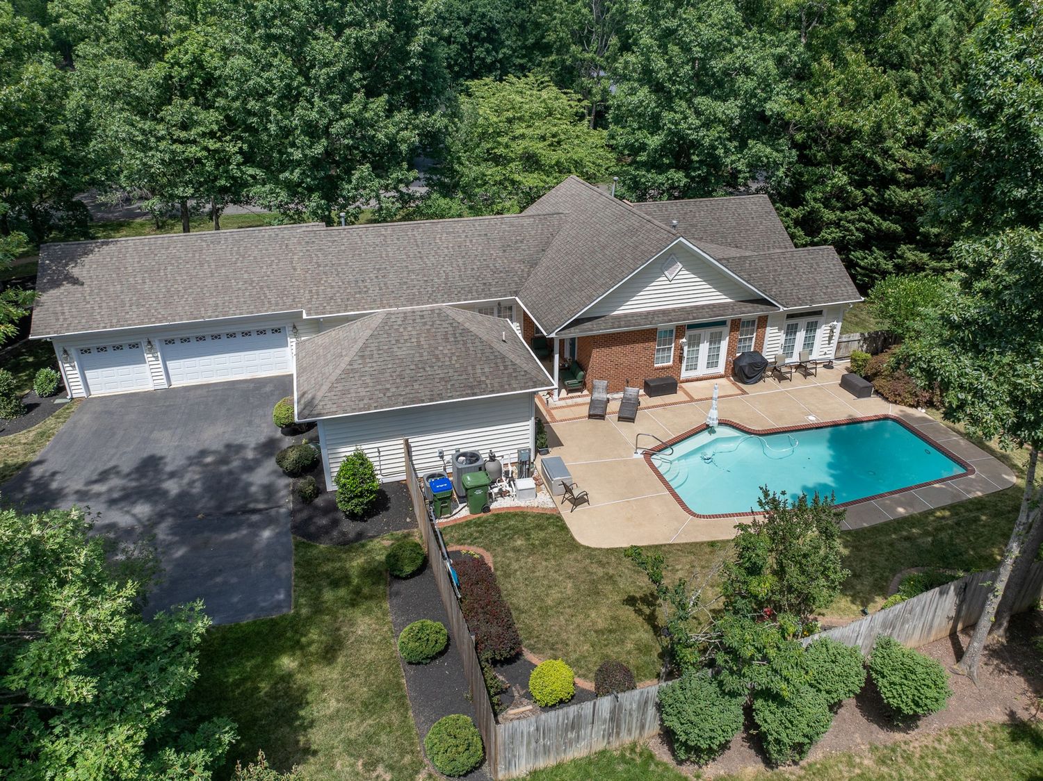 Aerial view of a brick ranch home with a backyard swimming pool, patio, three-car garage, and lush tree surroundings.