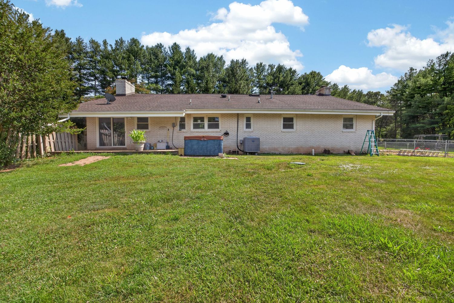 Ranch-style brick home with expansive green lawn, rear patio, hot tub, and tall pine trees in background on sunny day.