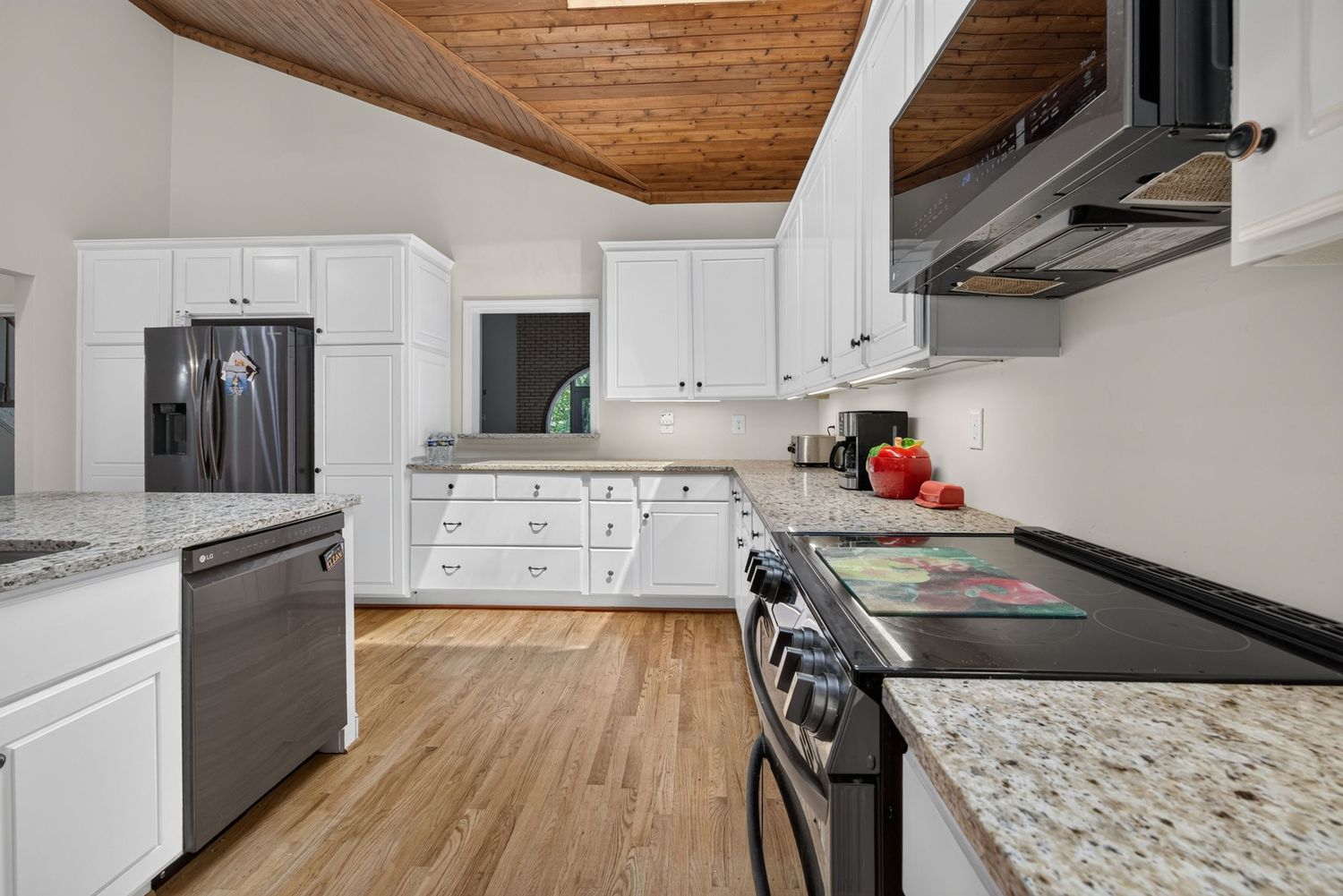 Modern kitchen with white cabinets, granite countertops, stainless steel appliances, and hardwood floors under a wood-paneled ceiling.