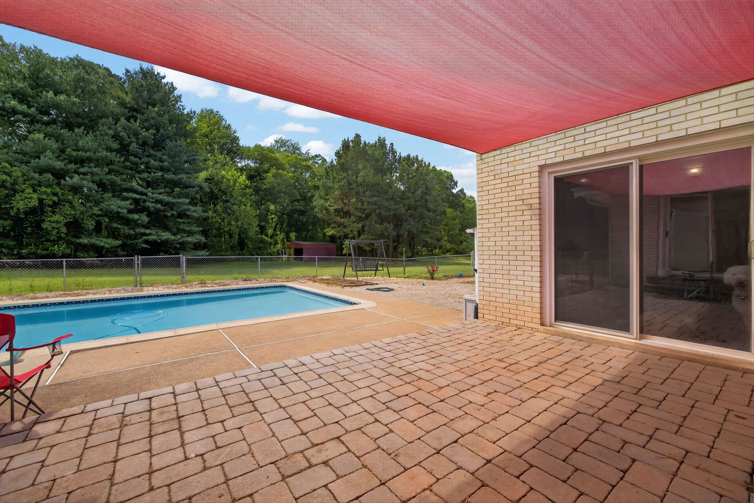 Brick patio with red shade sail overlooking an inground pool surrounded by lush green trees and fenced backyard.
