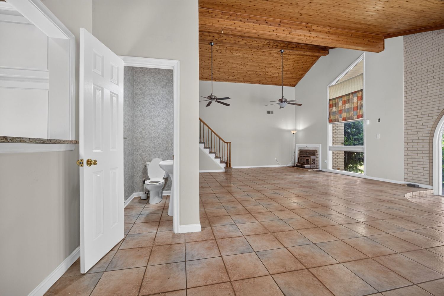 Spacious open-plan living area with tile floors, vaulted wood ceiling, fireplace, staircase, and adjacent bathroom.