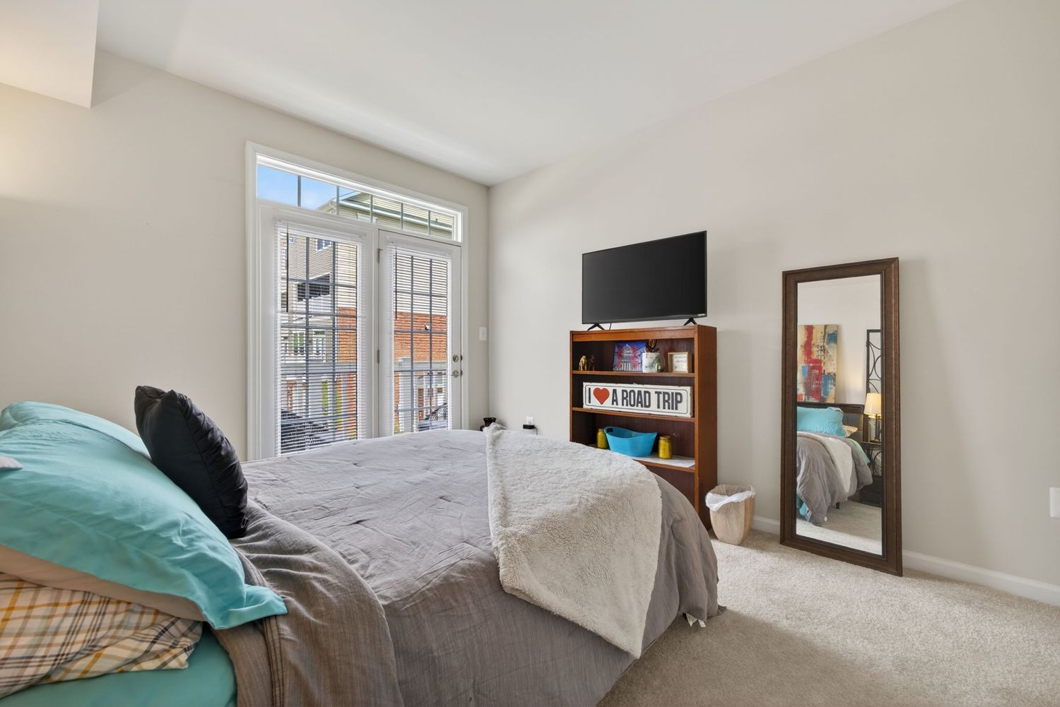 Bright bedroom with teal pillows, gray bedding, wall-mounted TV, bookshelf, floor mirror, and French doors to a balcony.