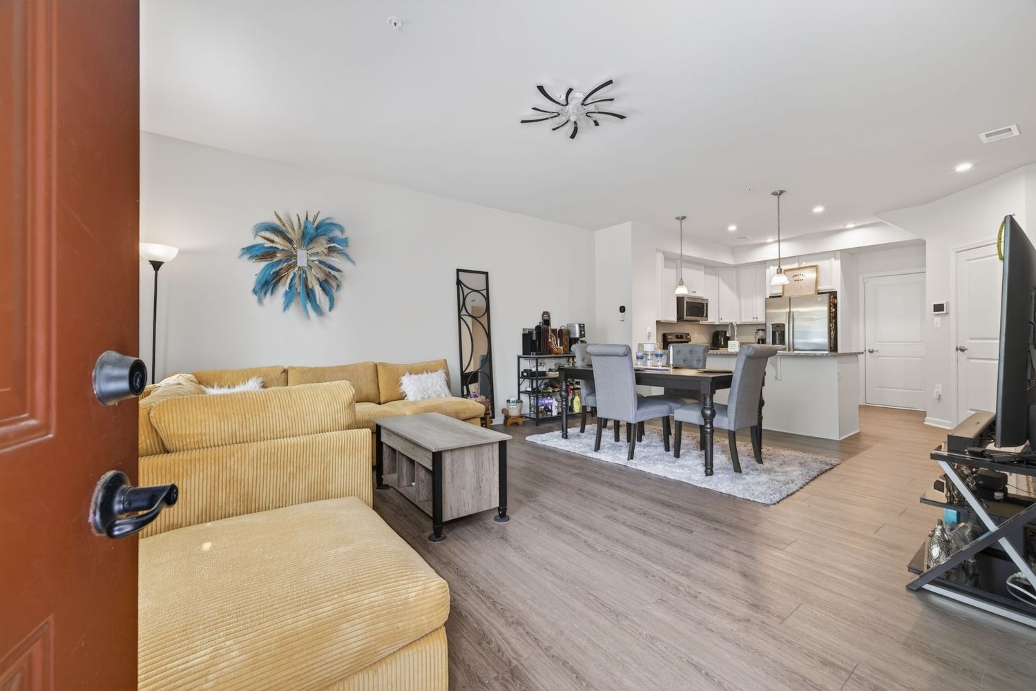 Open-concept living room with gold sectional sofa, wood floors, and modern kitchen with pendant lights in background.