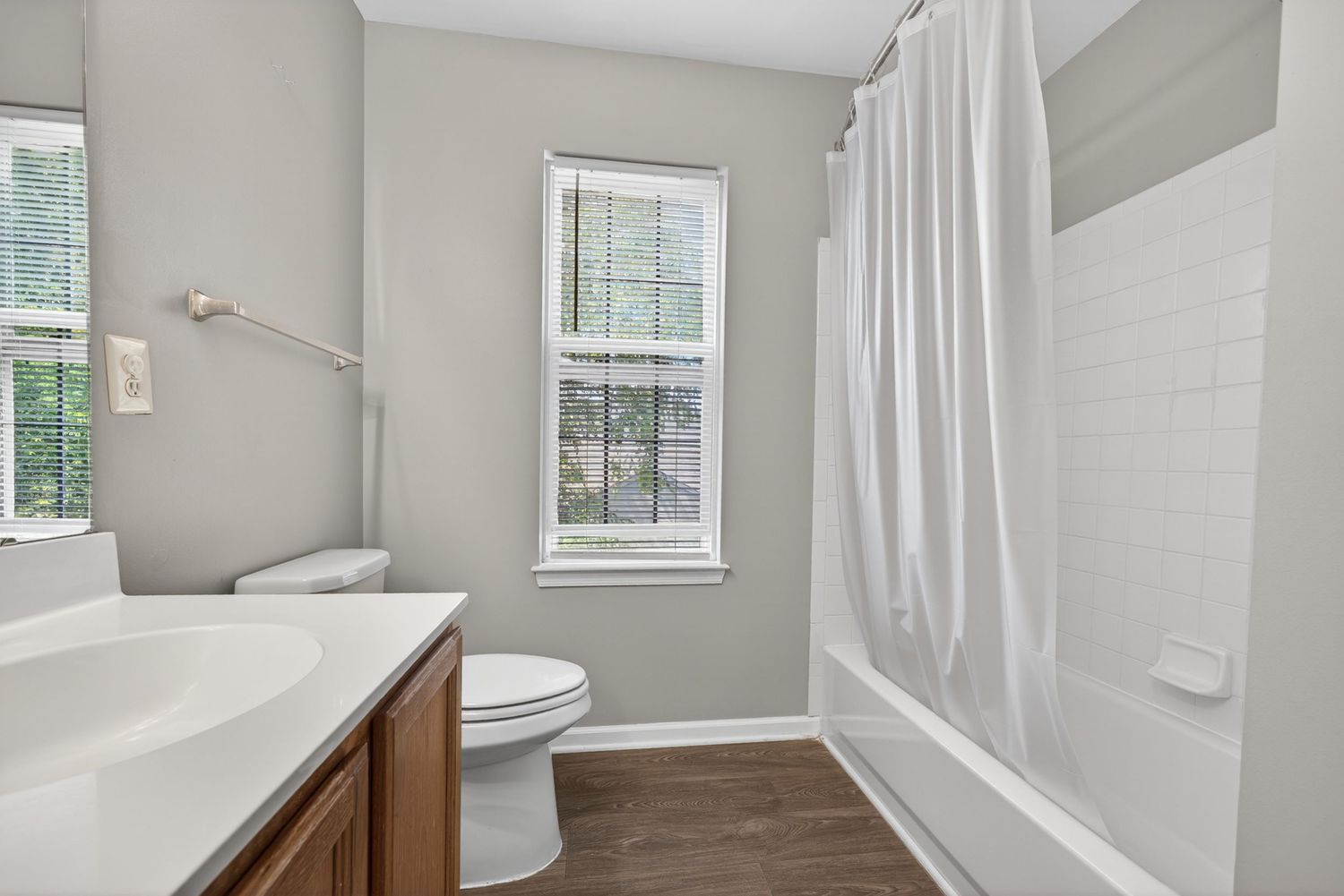 Clean bathroom with wood vanity, white tub with shower curtain, toilet, and two windows with natural light.