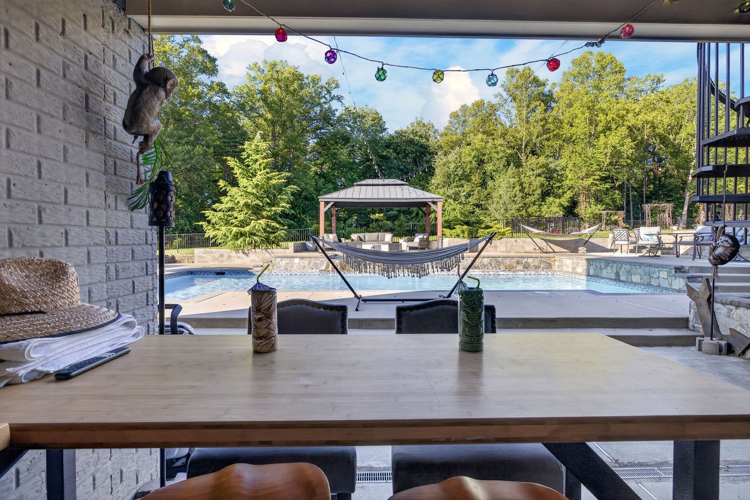 Outdoor patio dining area overlooking a backyard pool, gazebo, and lush trees with festive string lights overhead.