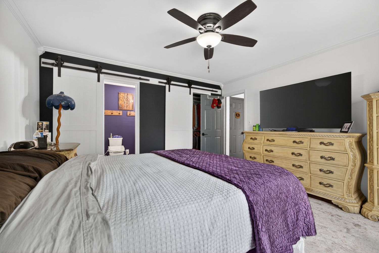 Bedroom with ceiling fan, barn sliding doors, large TV on dresser, and a bed with purple accent blanket.