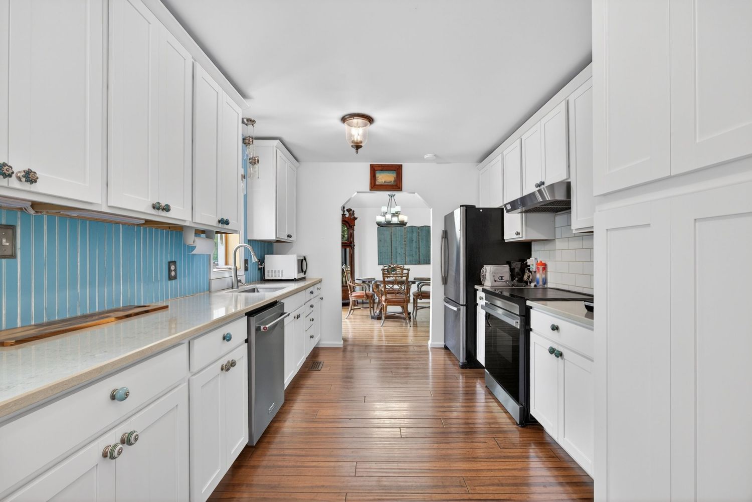 Bright galley kitchen with white cabinets, hardwood floors, blue tile backsplash, and stainless steel appliances.